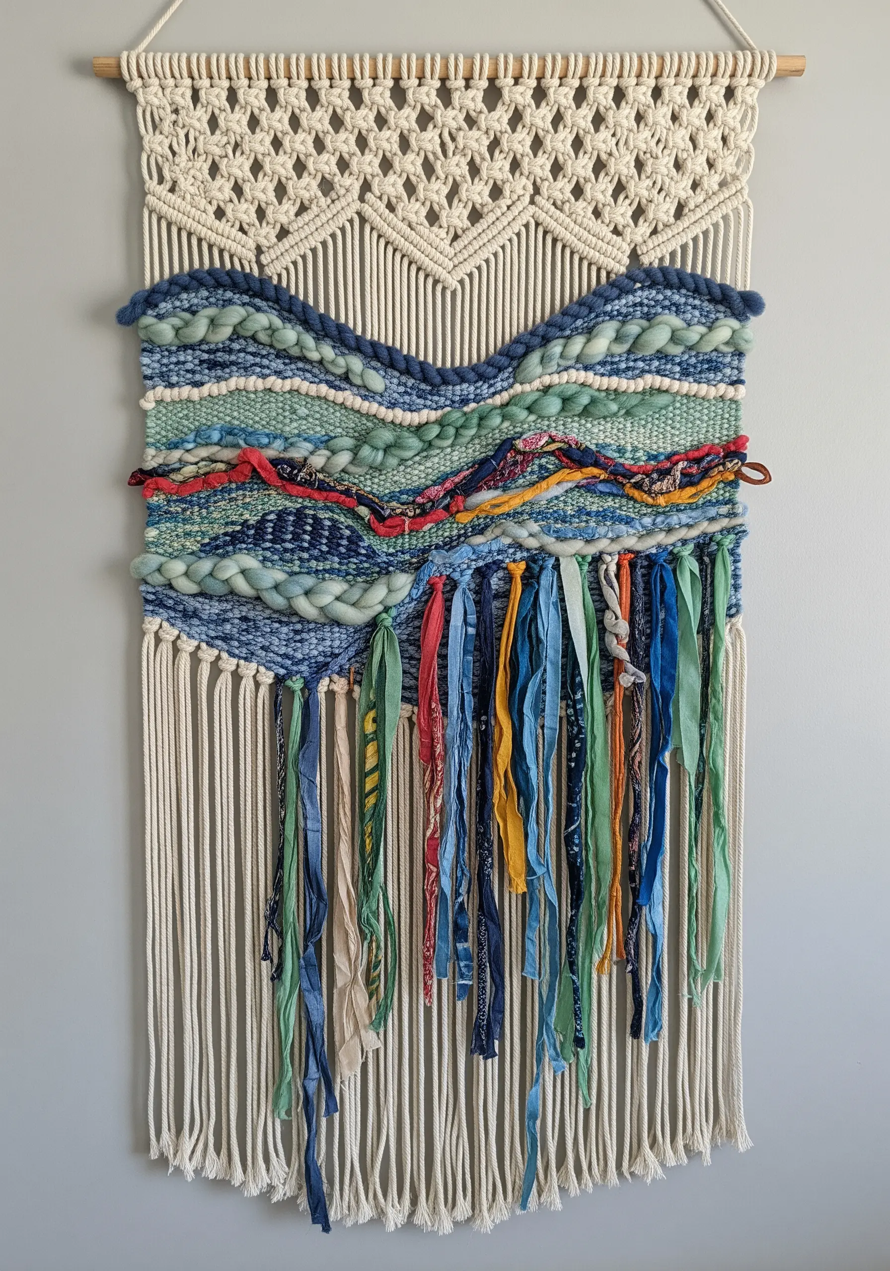 Macrame wall hanging with a central woven section featuring blues, greens, and fabric strips.