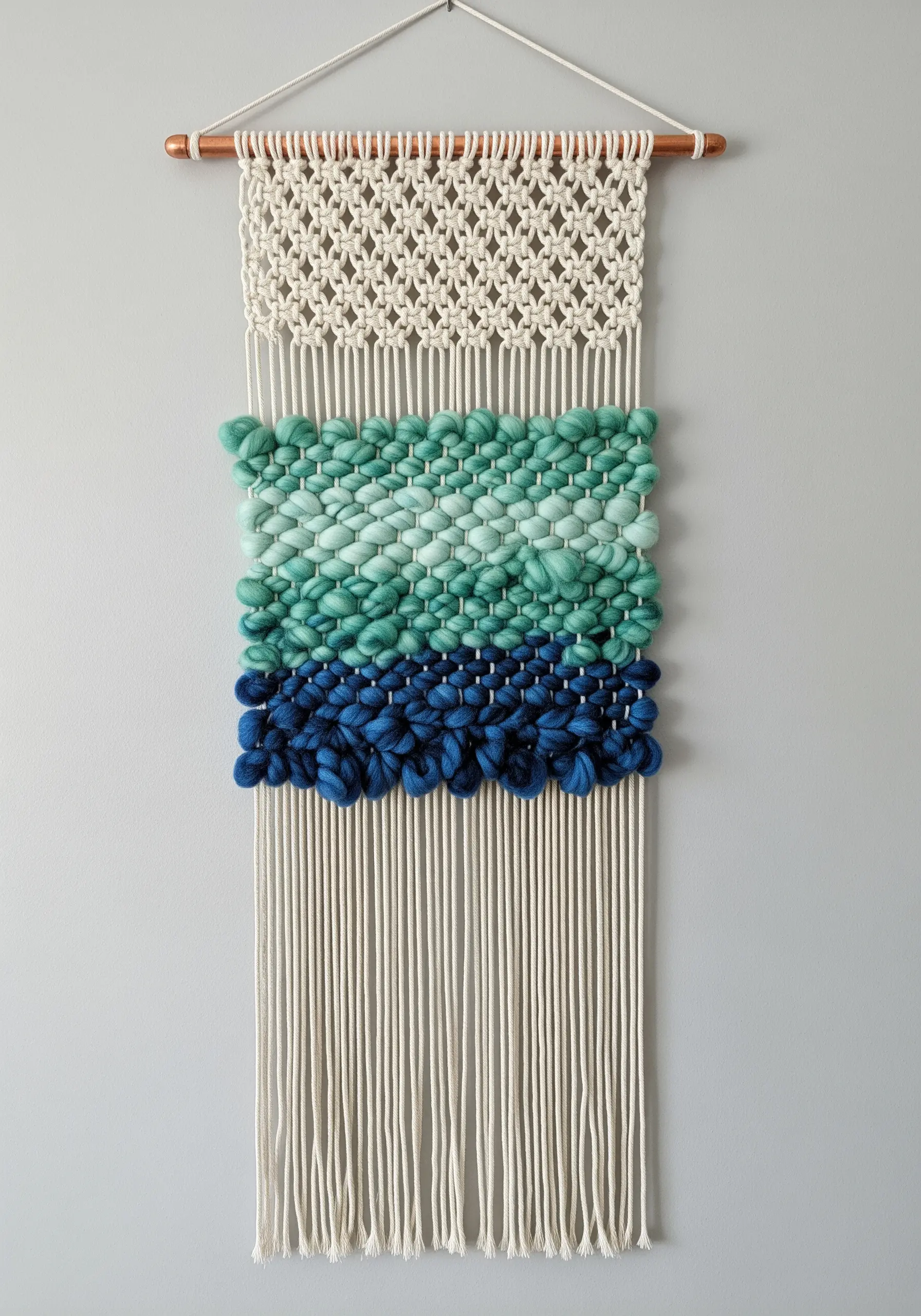 A wall hanging combining macrame knots with a woven section in shades of blue and green.