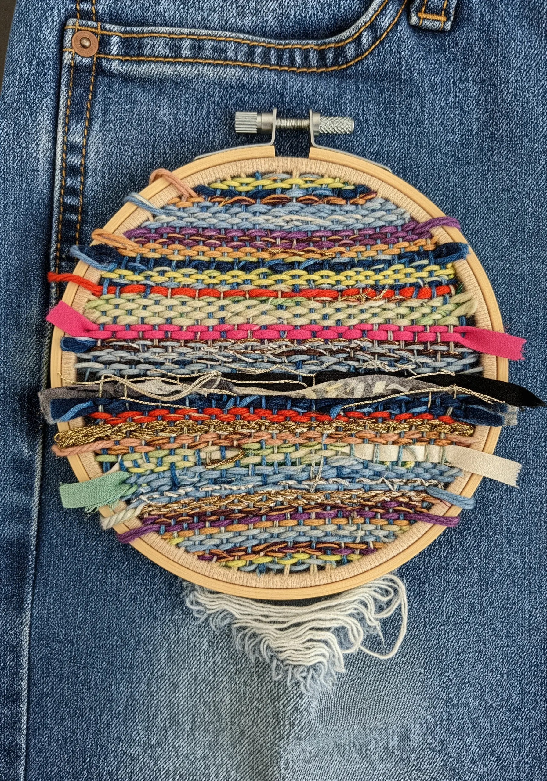 A small embroidery hoop used as a loom to weave colorful fabric scraps and yarns.