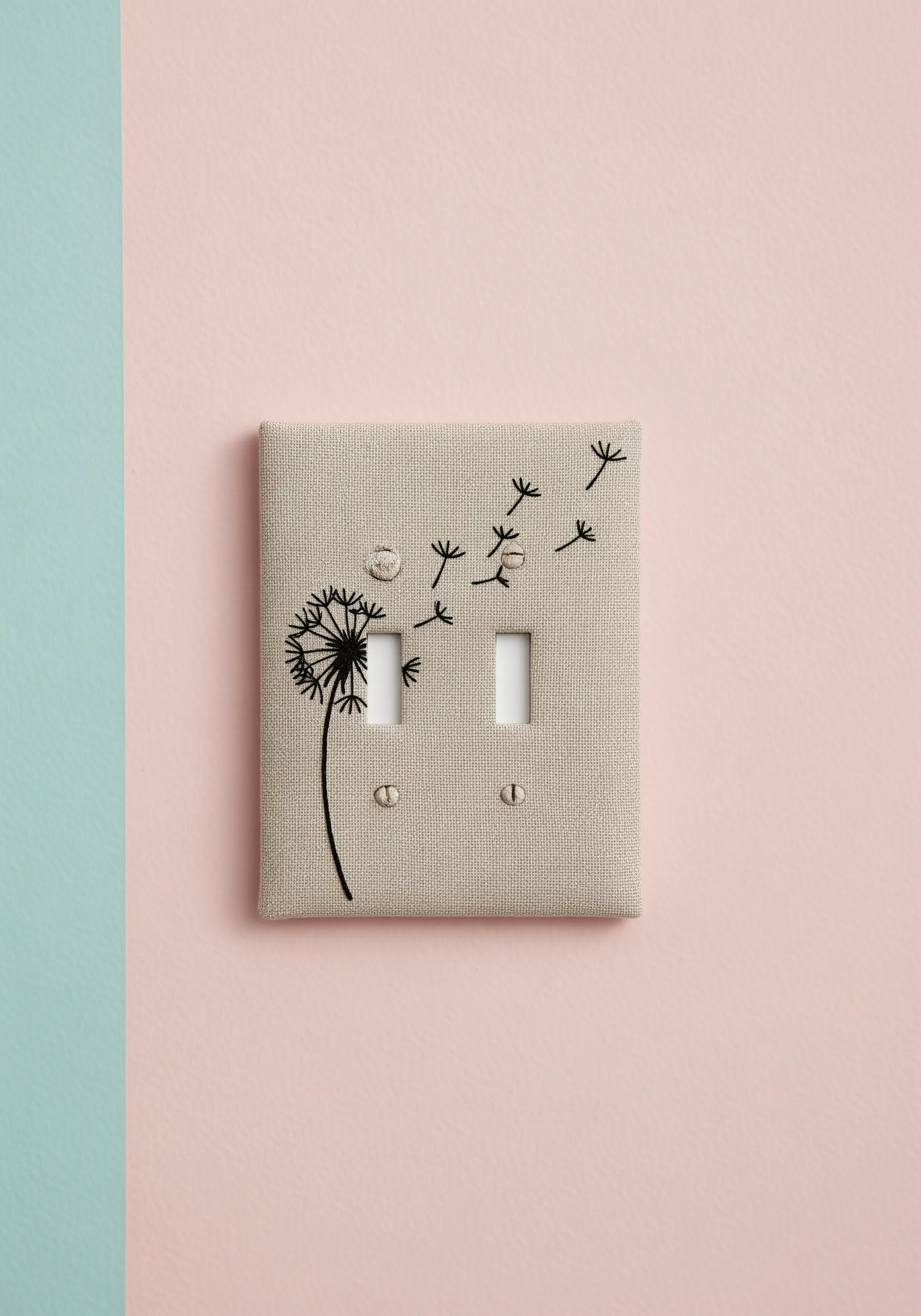 A linen-covered light switch plate embroidered with a black dandelion silhouette.