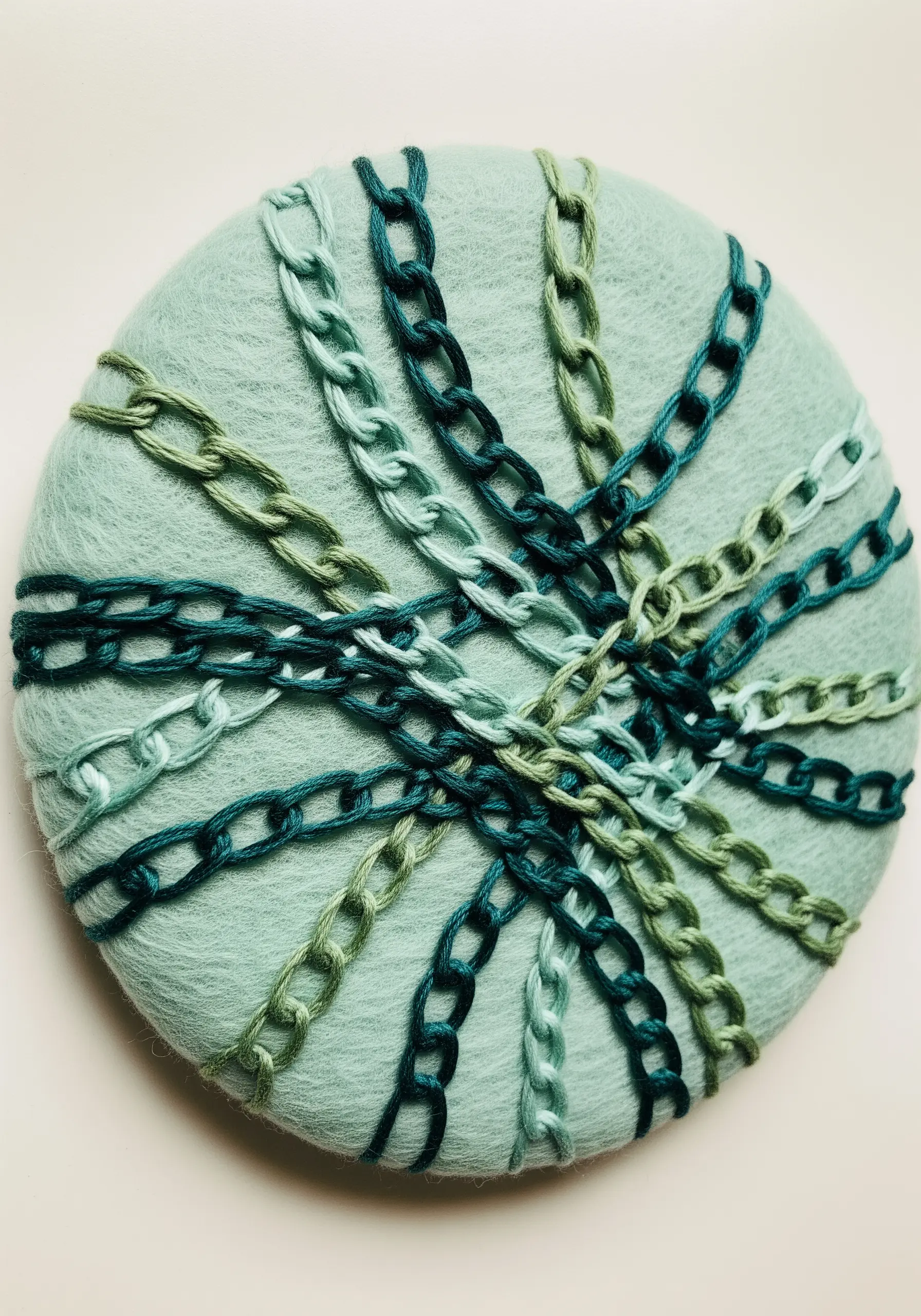 Interlocking lines of green and teal chain stitch on a round, light green felt base.