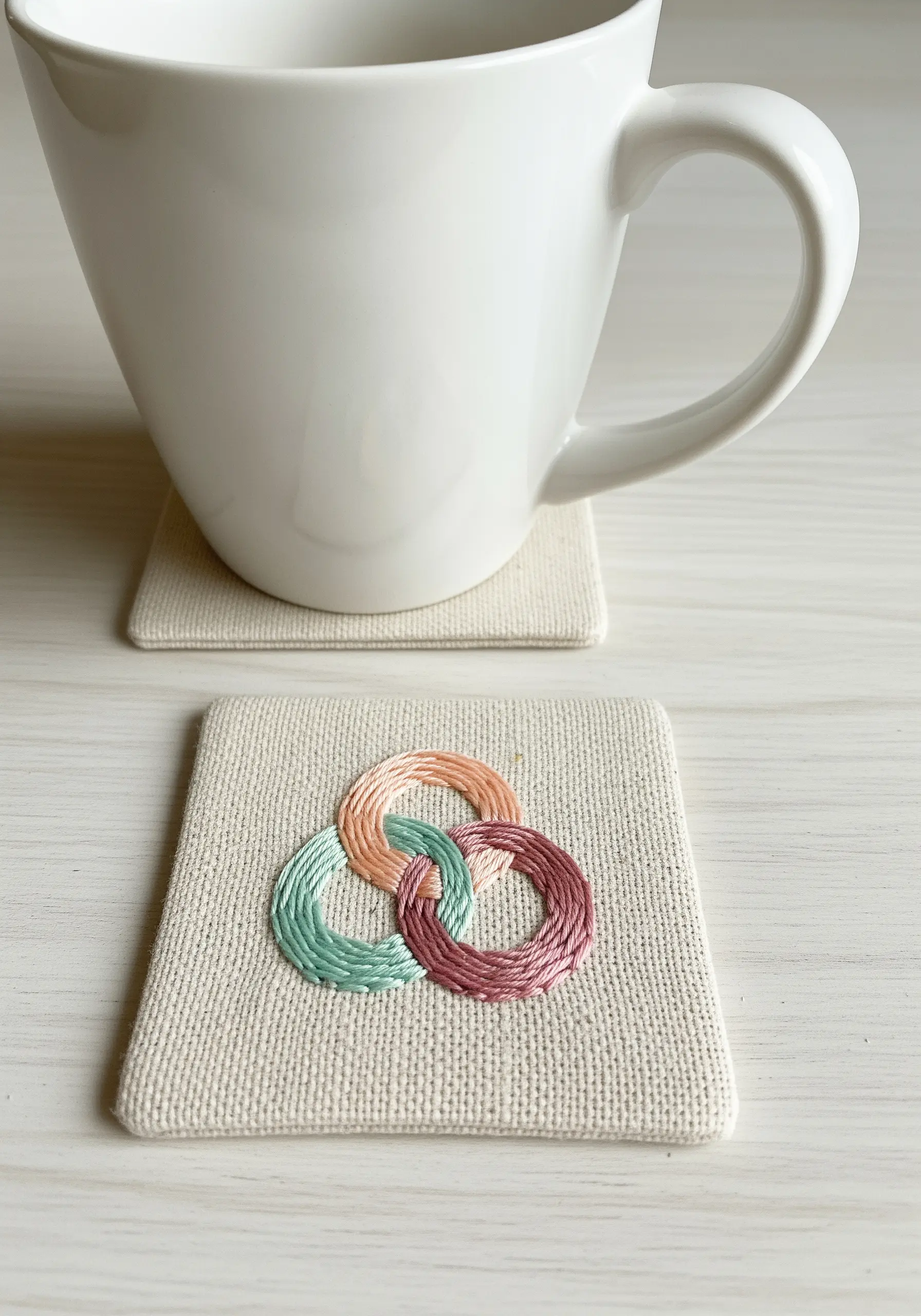 Three interlocking rings in teal, pink, and mauve embroidered on a fabric coaster.