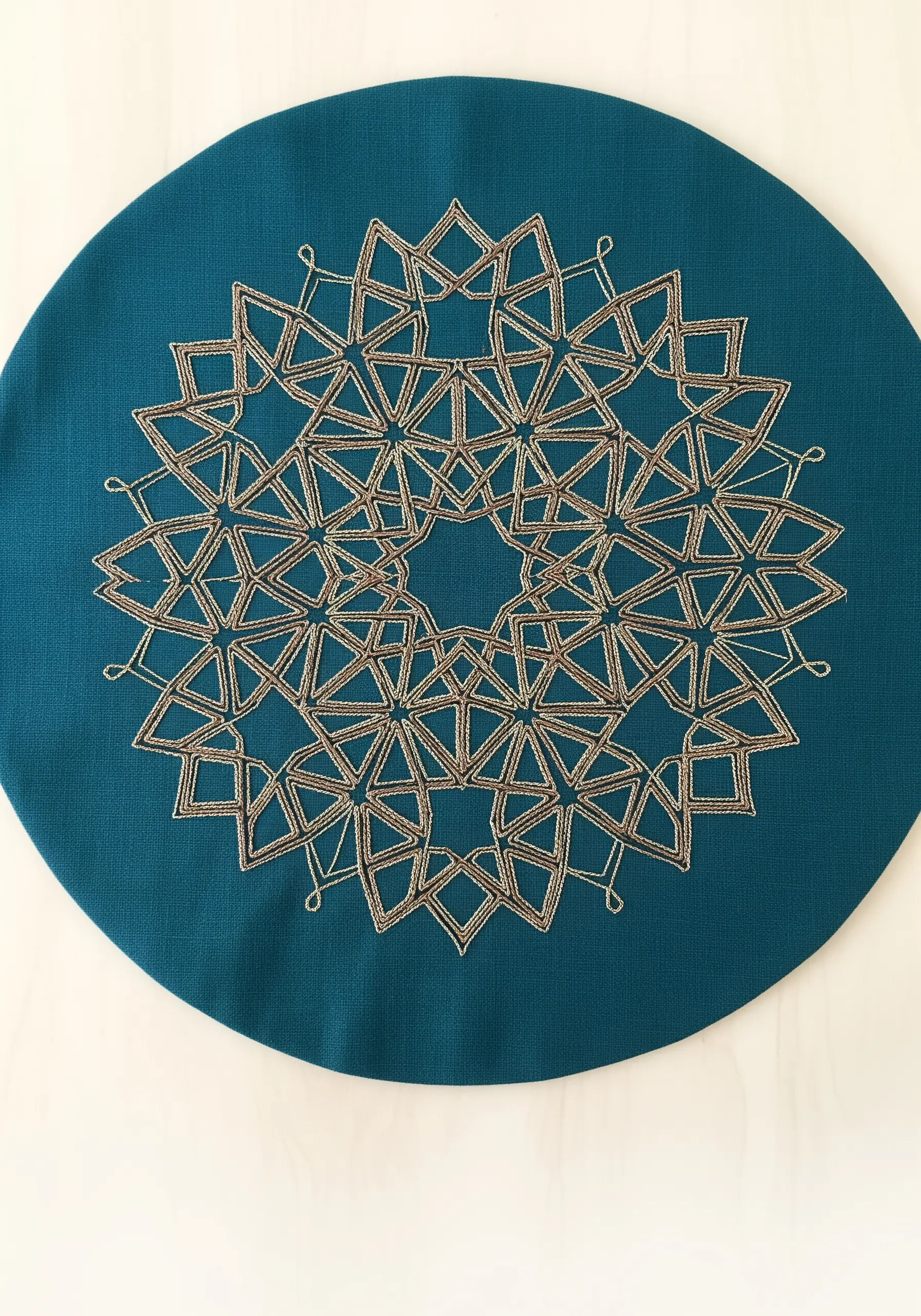 A geometric Islamic star mandala stitched in gold metallic thread on a teal placemat.