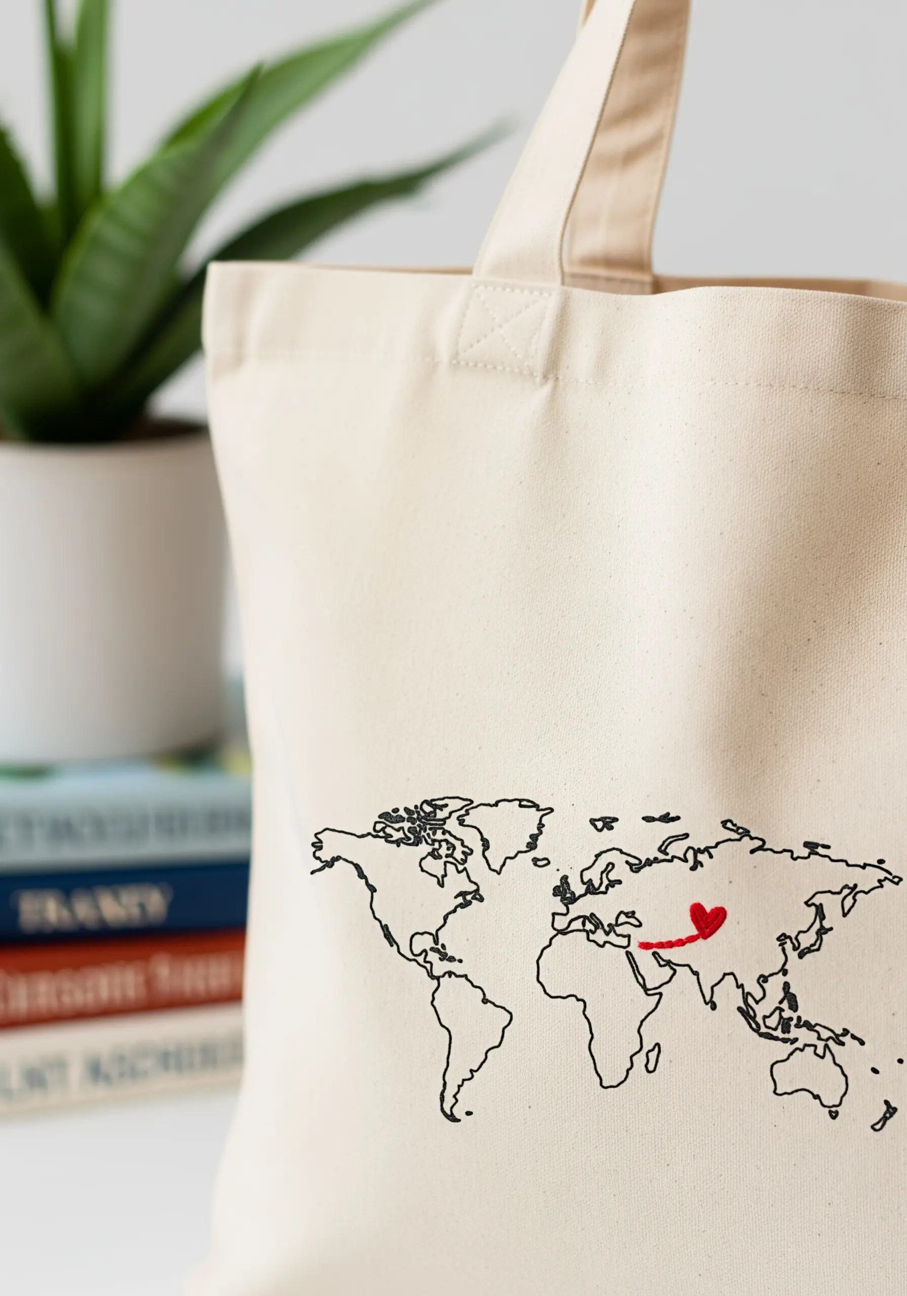 Minimalist world map outline embroidered on a canvas tote bag, with a red heart over one region.