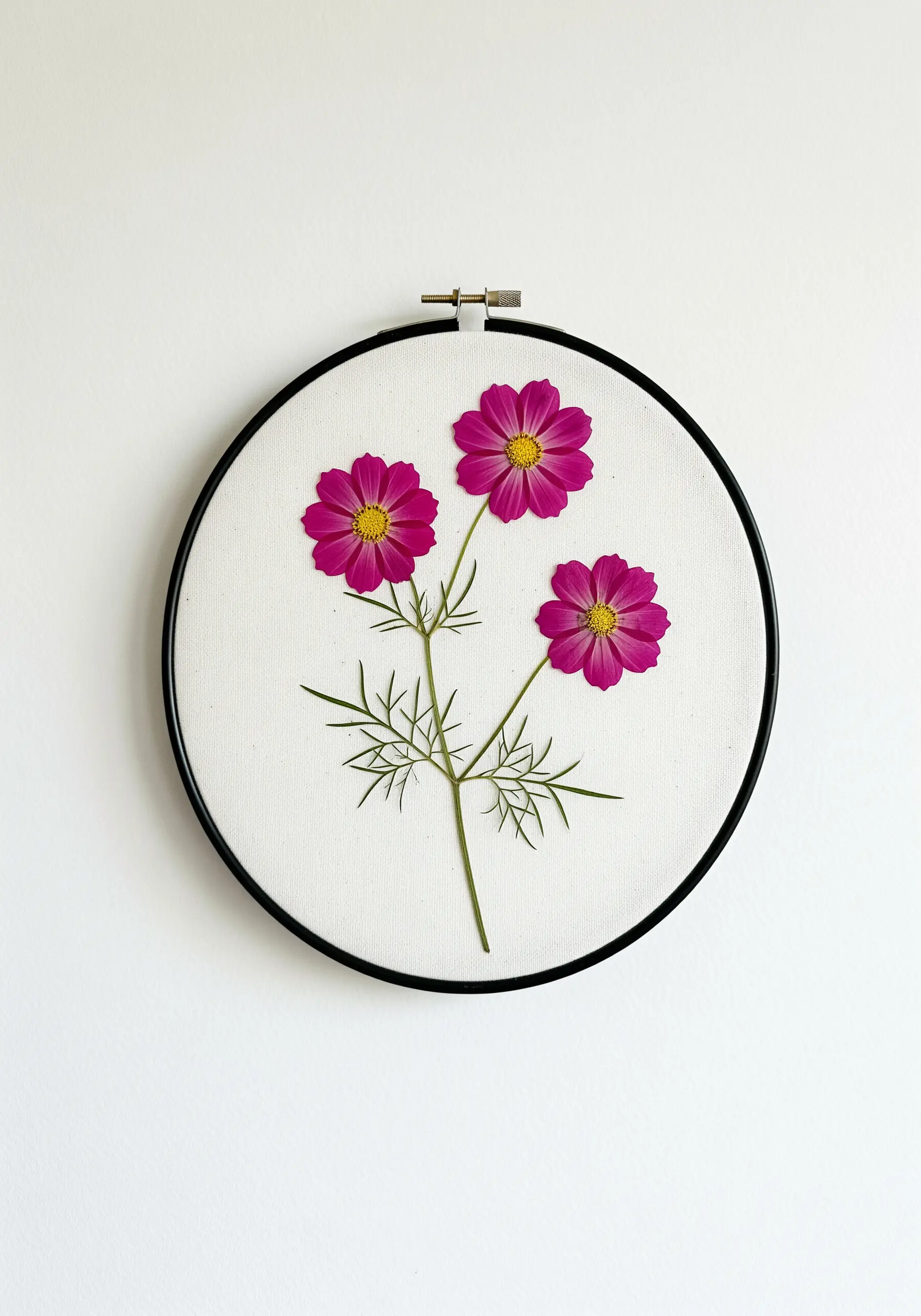 Three pressed pink cosmos flowers with delicate leaves stitched in a black embroidery hoop.