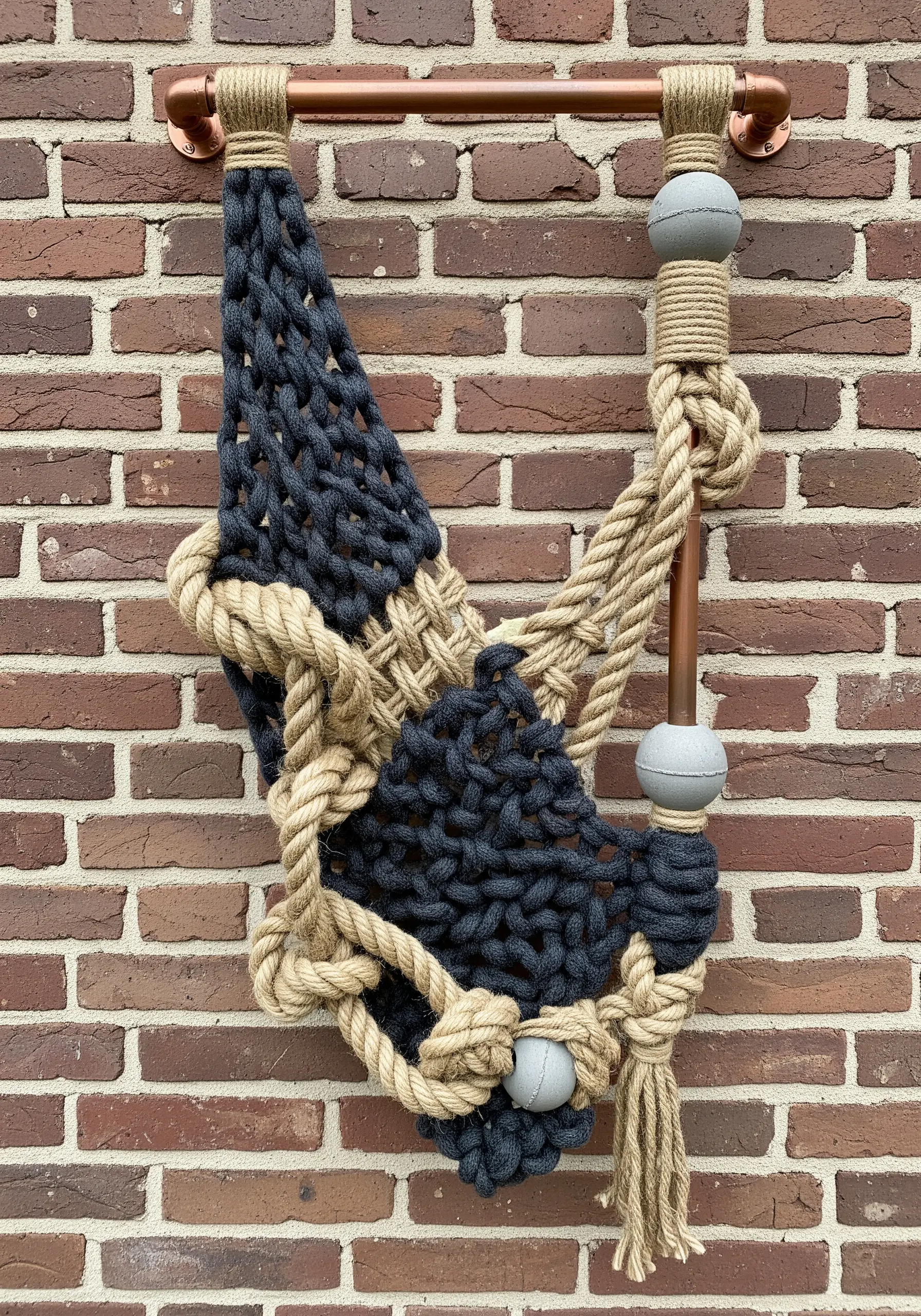 A sculptural wall hanging made of thick jute rope and chunky charcoal yarn, mounted on a copper pipe.