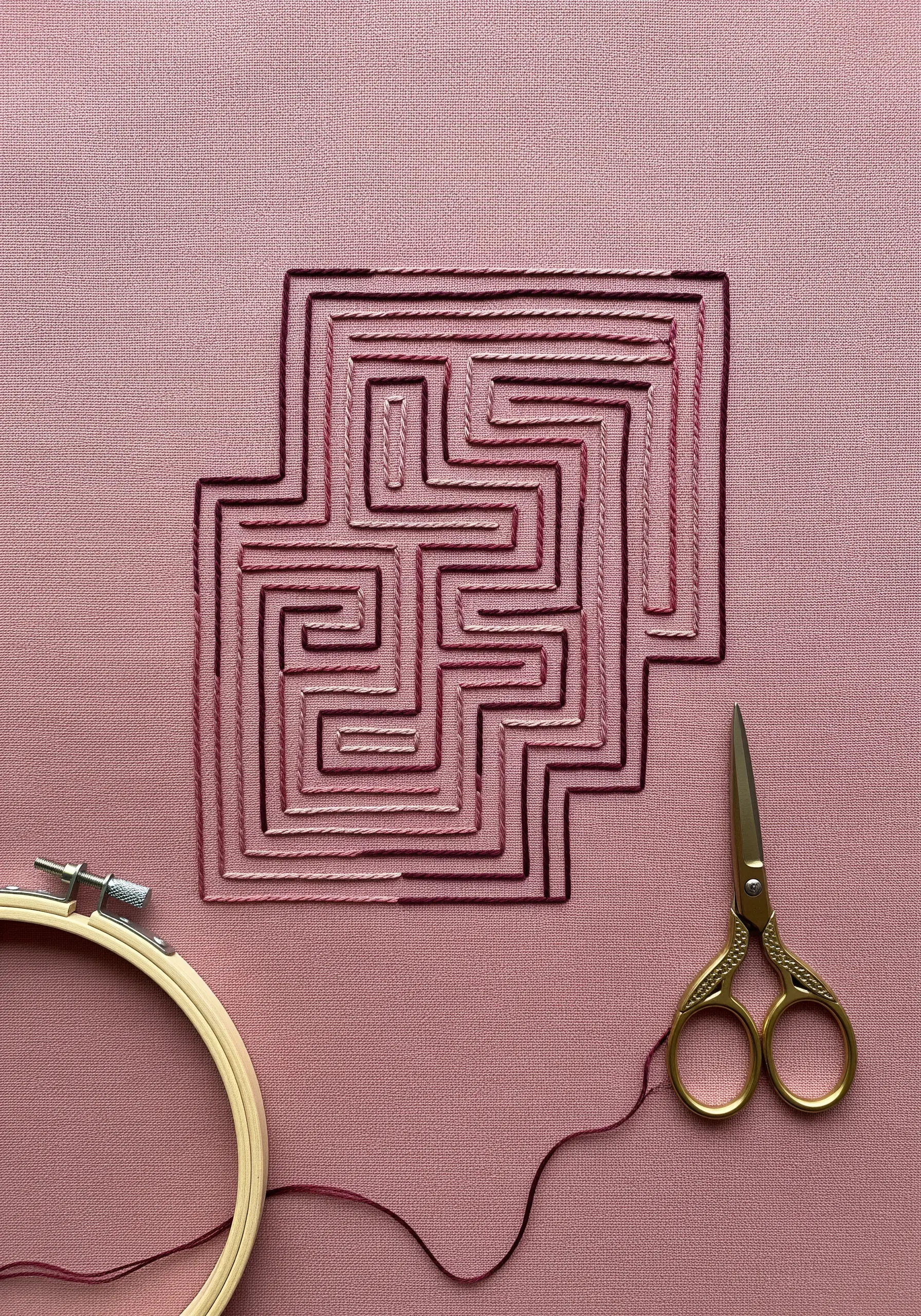 A geometric maze embroidered with variegated red-purple thread on pink fabric.