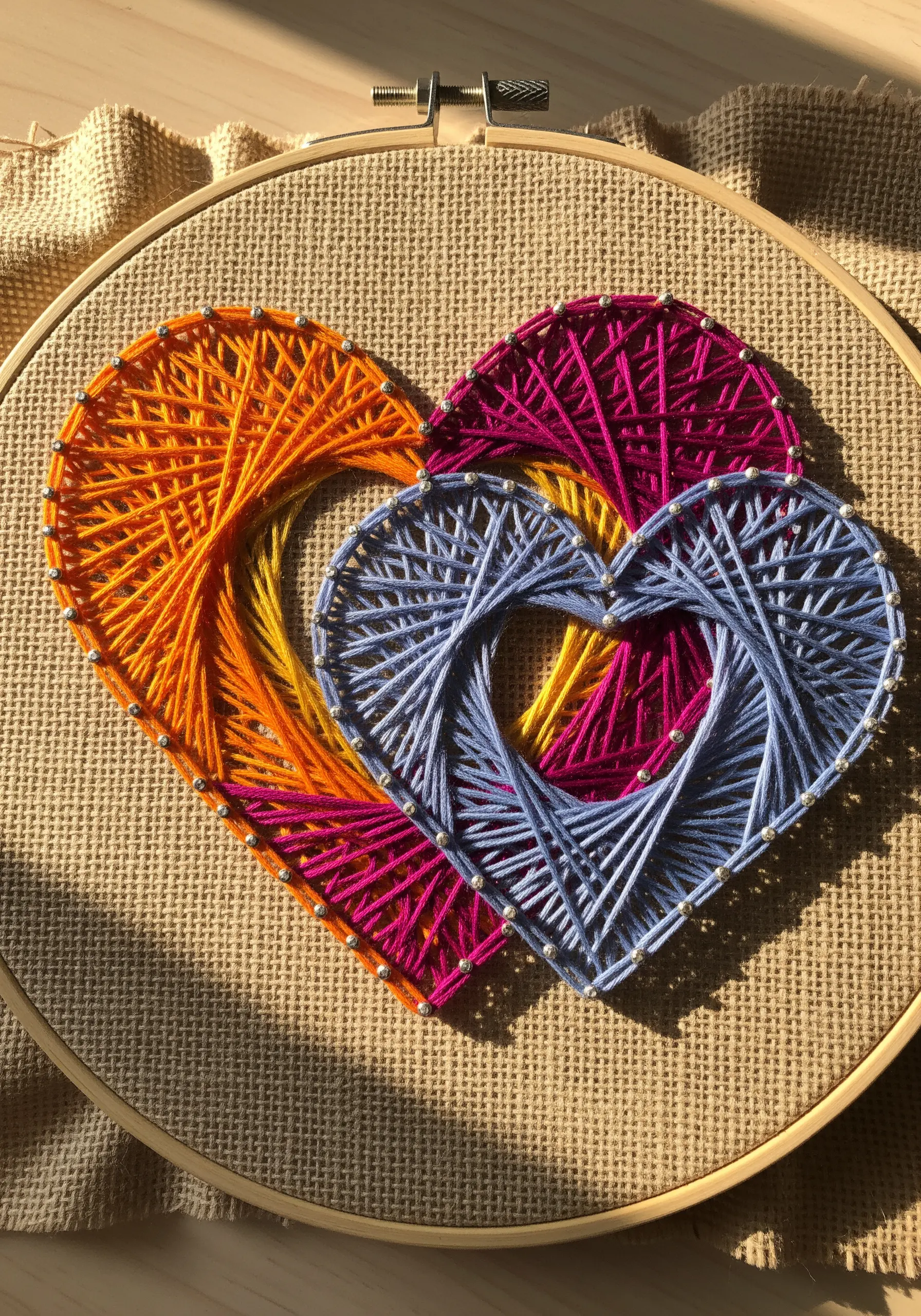 Two interlocking string art hearts in bright orange, magenta, and blue on coarse burlap fabric.