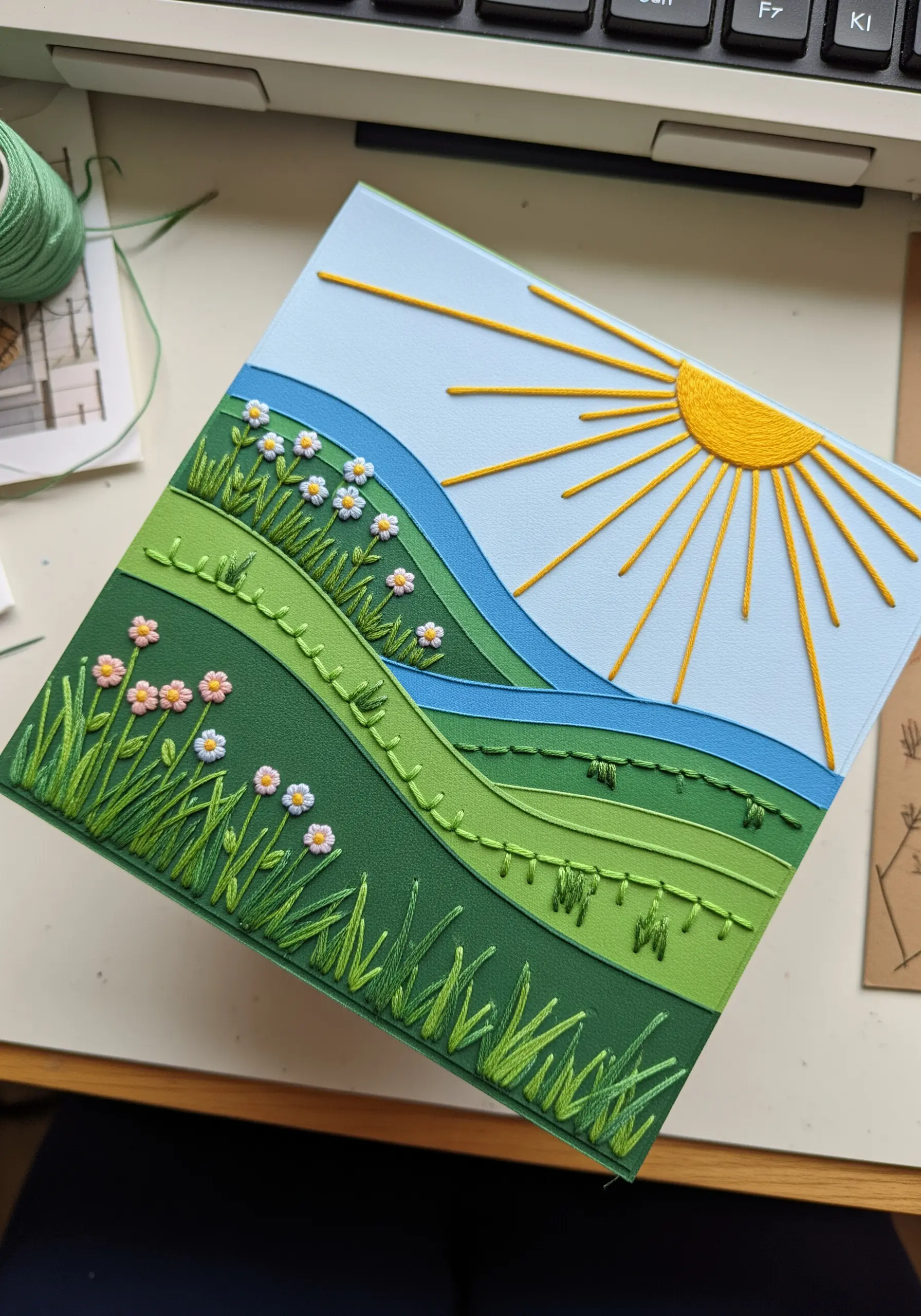 A layered paper landscape with embroidered grass, flowers, and sun details.