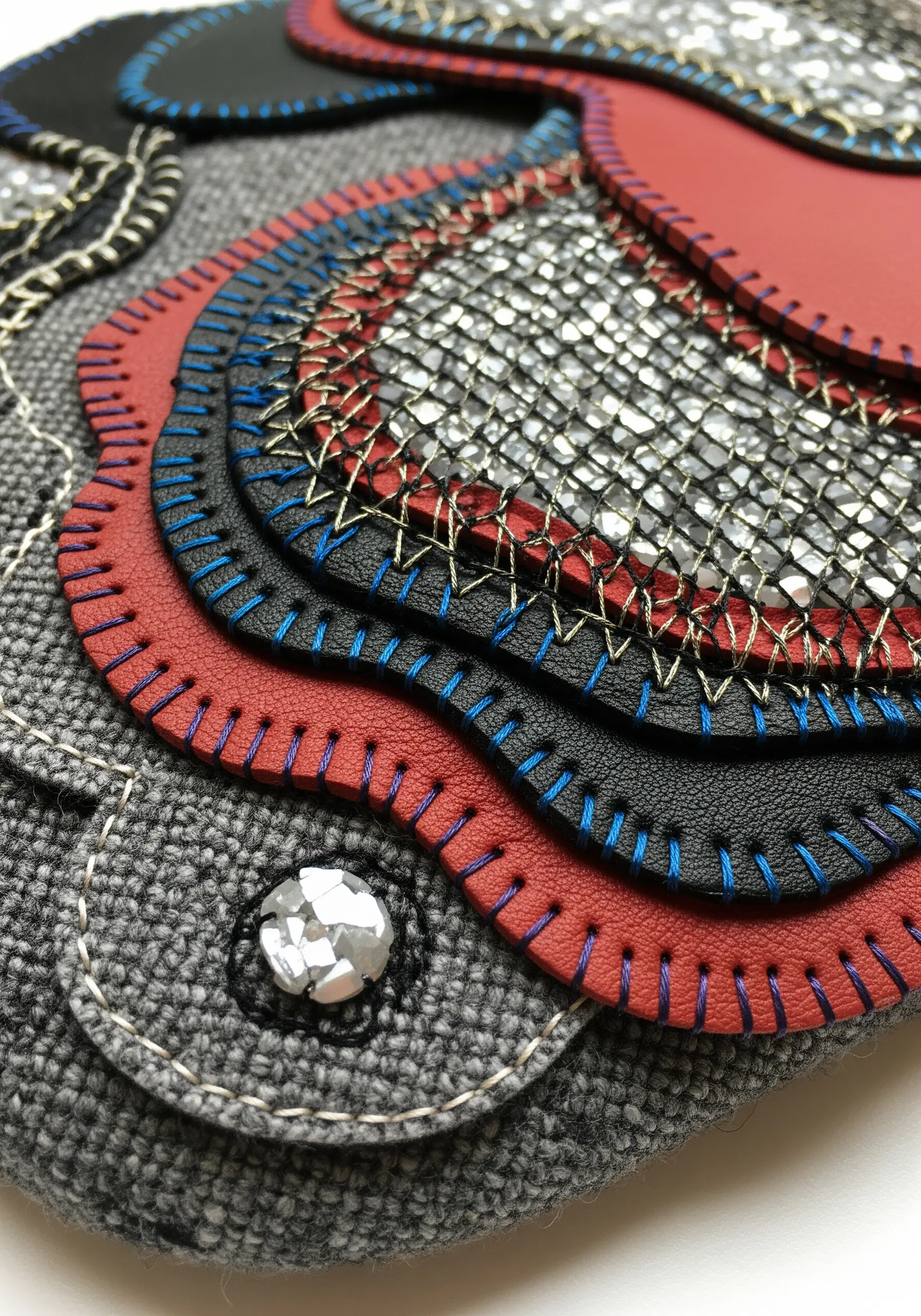 Layered faux leather and metallic mesh stitched together with colorful thread on a clutch.