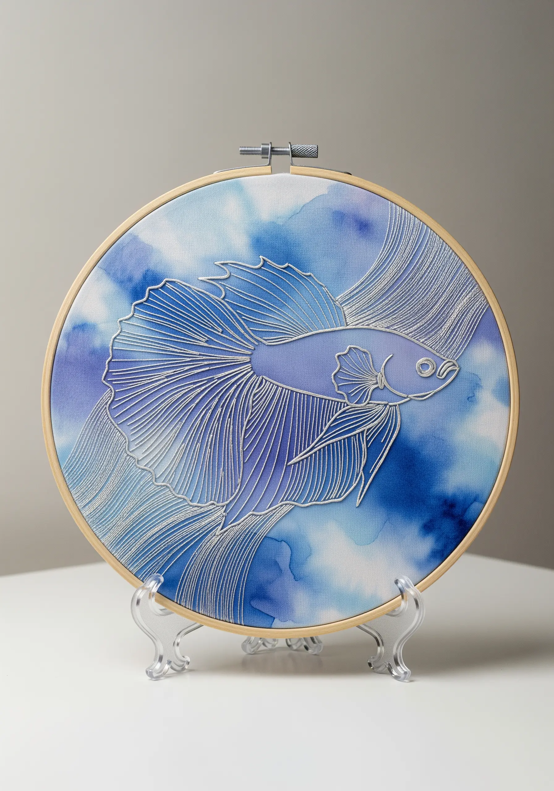 A white-thread betta fish embroidered over a blue watercolor background in a hoop.