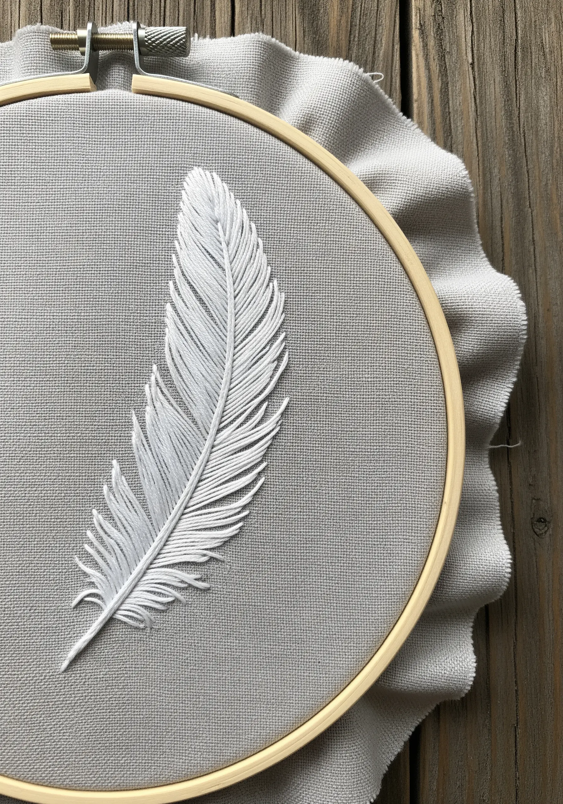 A delicate white feather embroidered on grey fabric using a fishbone stitch.
