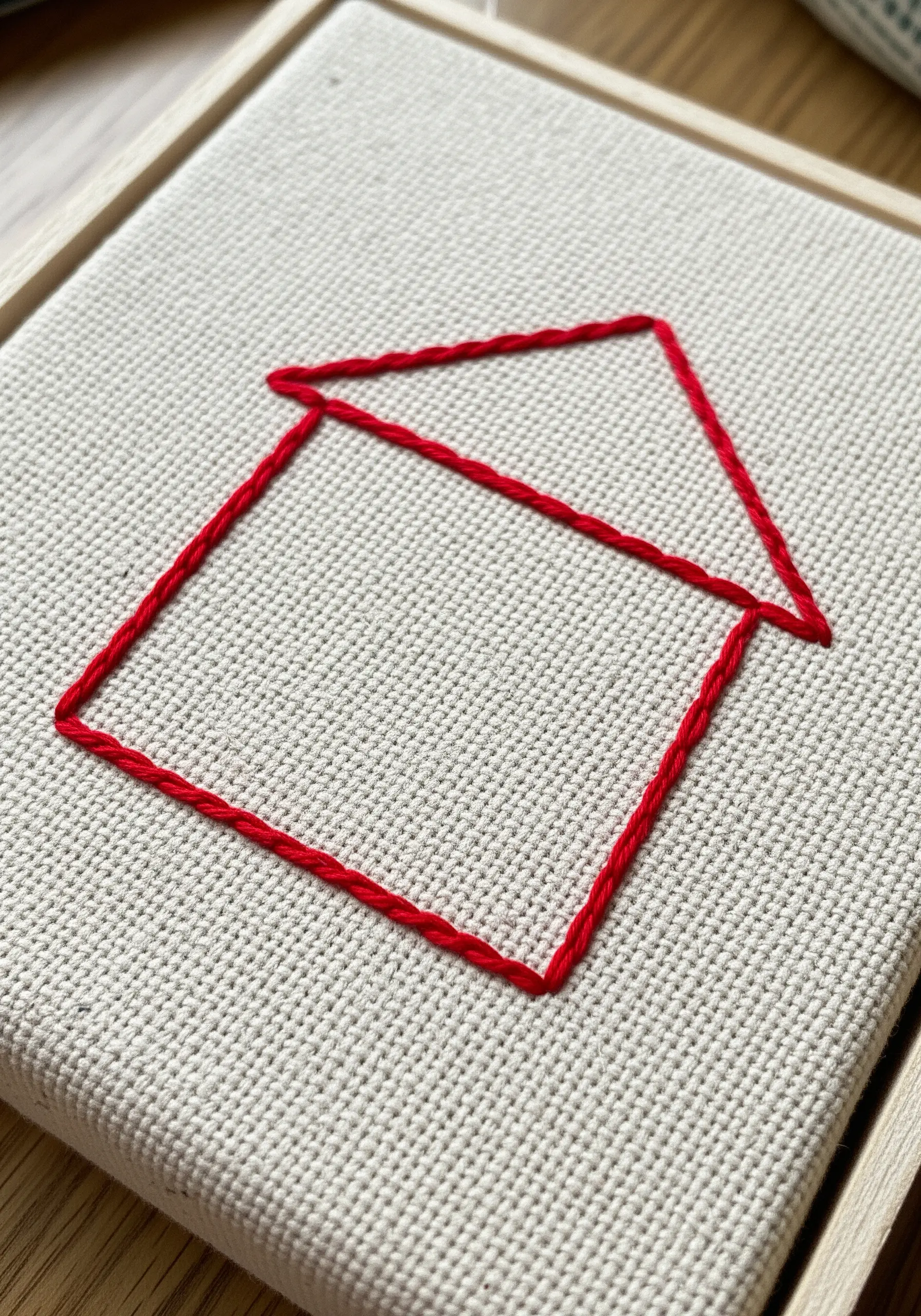 A simple red outline of a house embroidered on off-white Aida cloth in a frame.