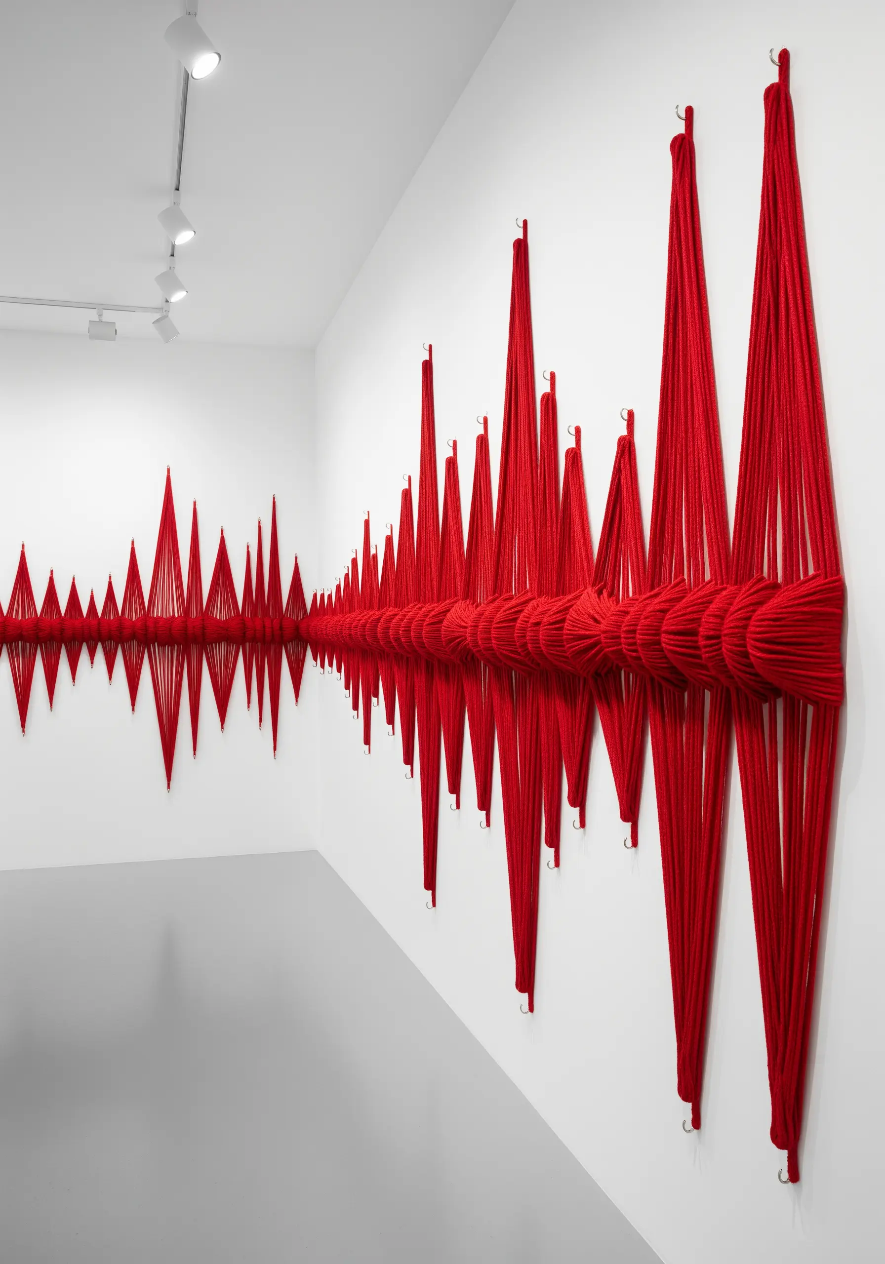 A red yarn installation on a white wall depicting a dramatic, rhythmic soundwave.