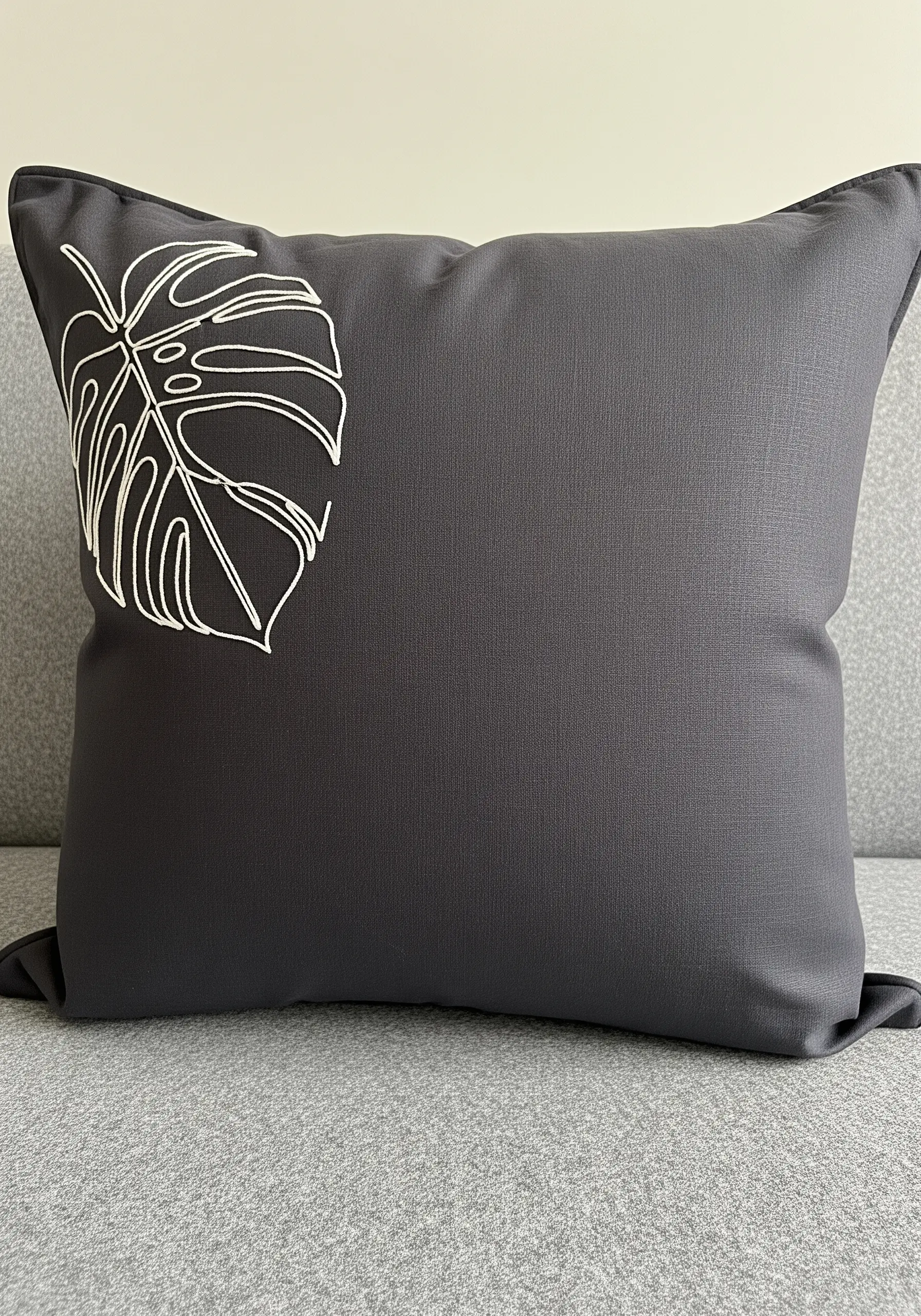 White line art embroidery of a monstera leaf in the top corner of a dark grey cushion.