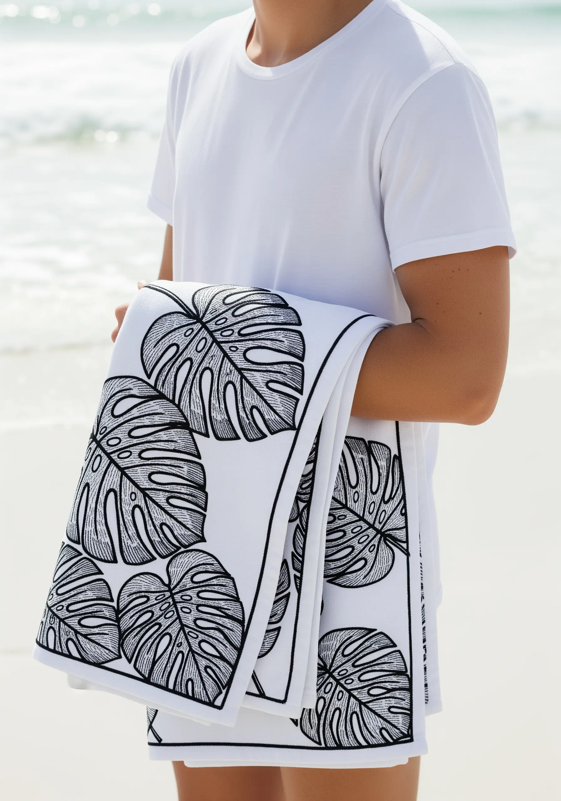 A bold black line-art pattern of monstera leaves embroidered on a white beach towel.