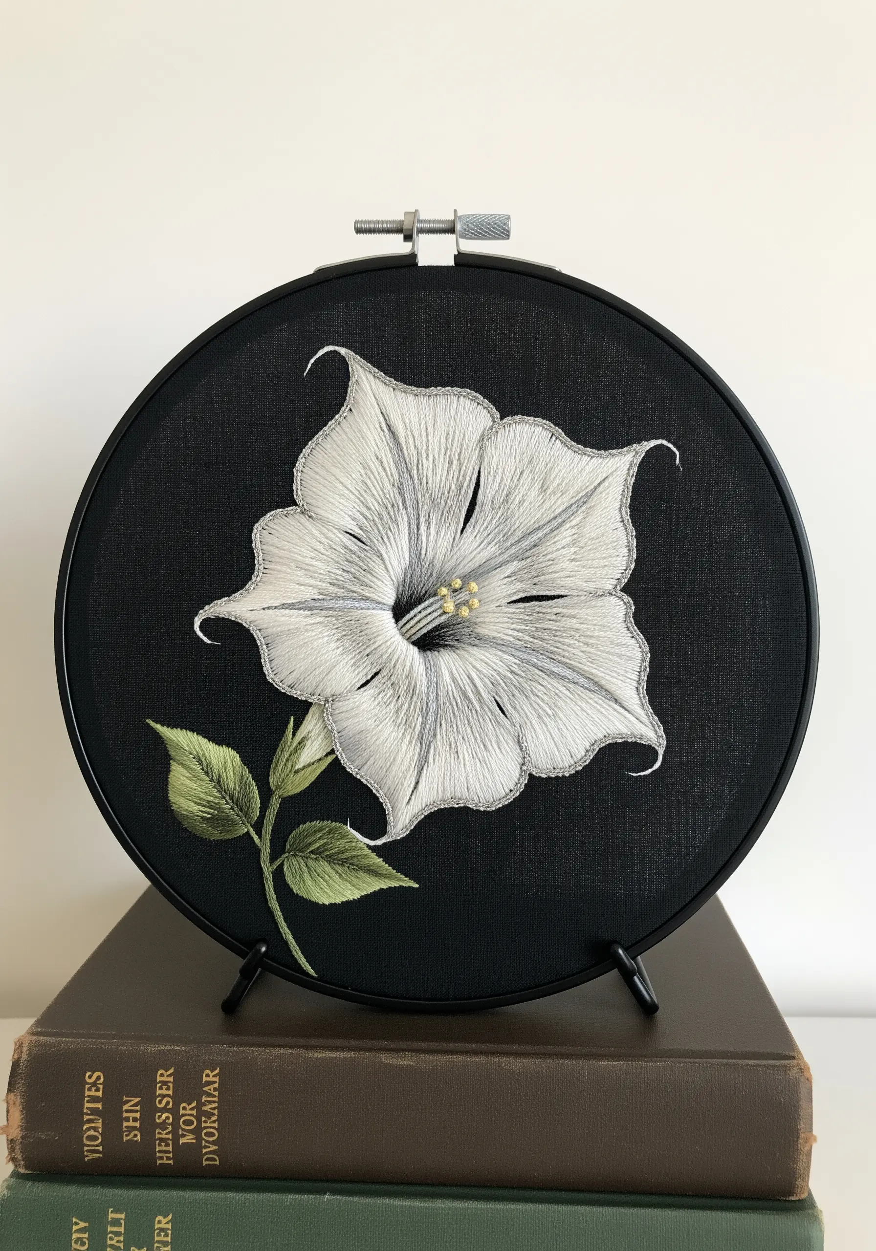 Thread-painted white datura flower with grey shading on black fabric in a black hoop.