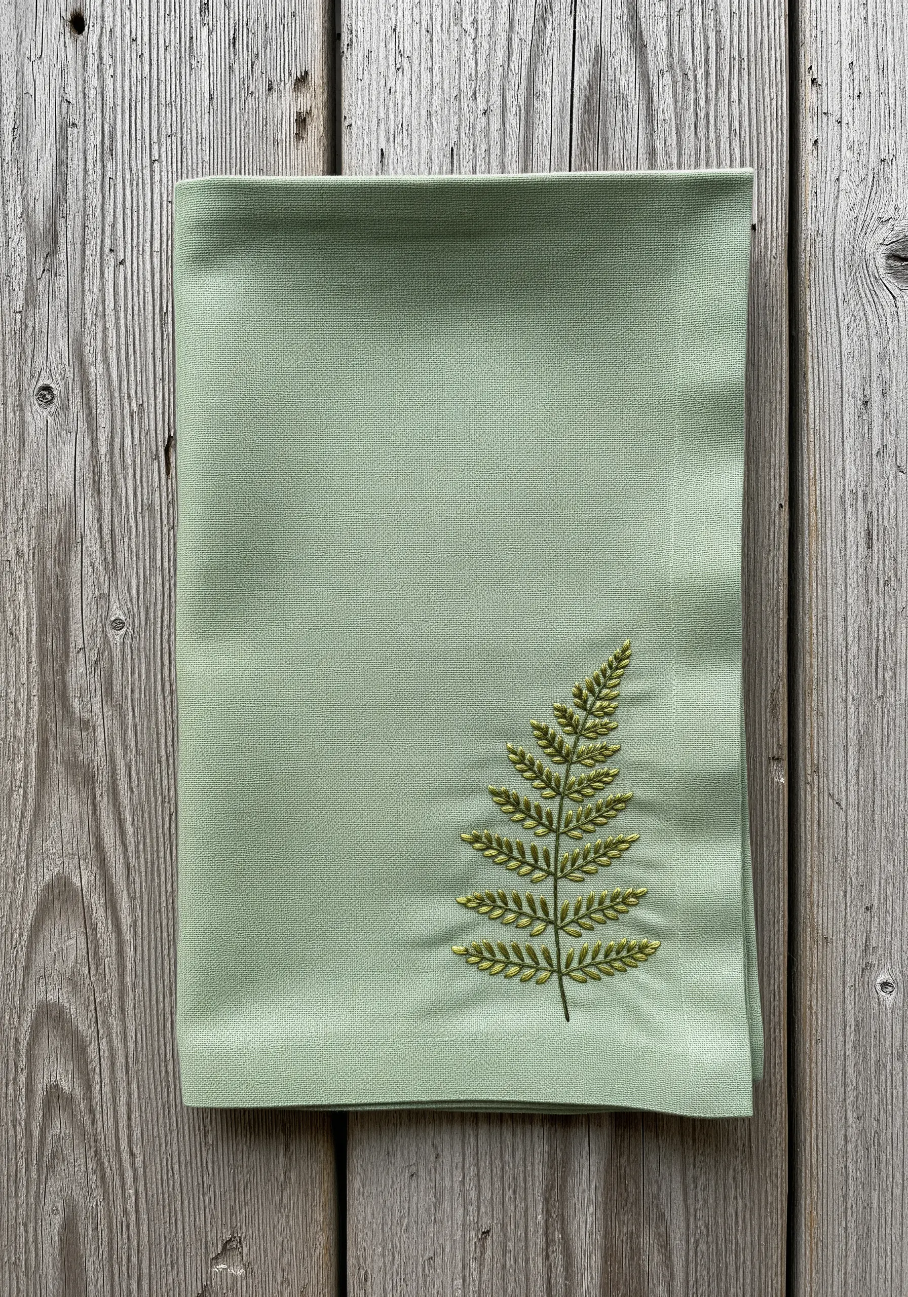 A single fern leaf embroidered in green on a sage green linen napkin.