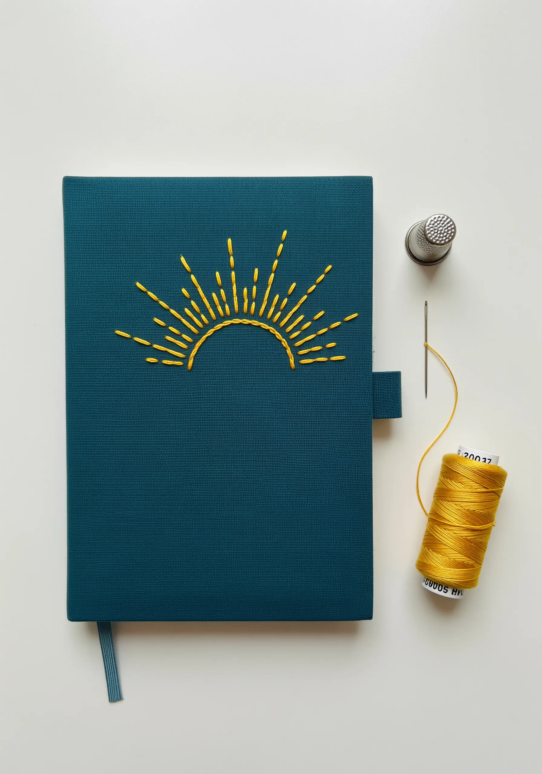 A simple yellow sunburst motif embroidered on the cover of a teal journal.