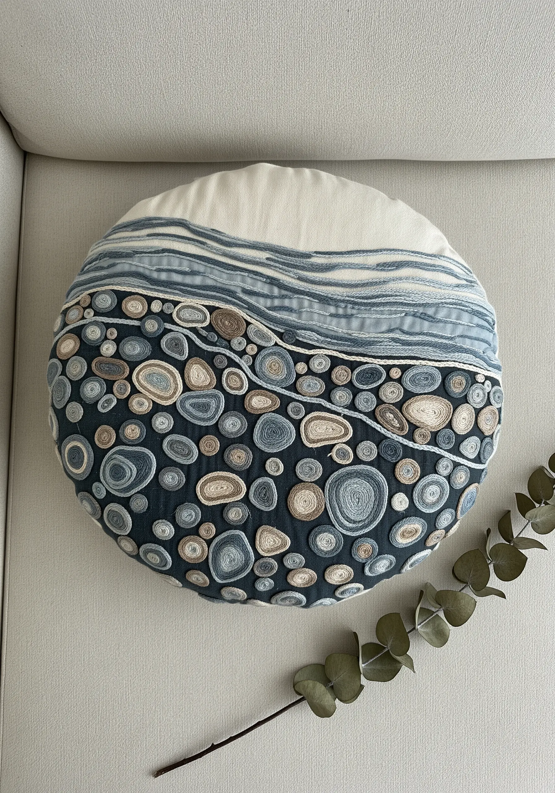 Round cushion decorated with coiled yarn appliqué in shades of blue and neutral.