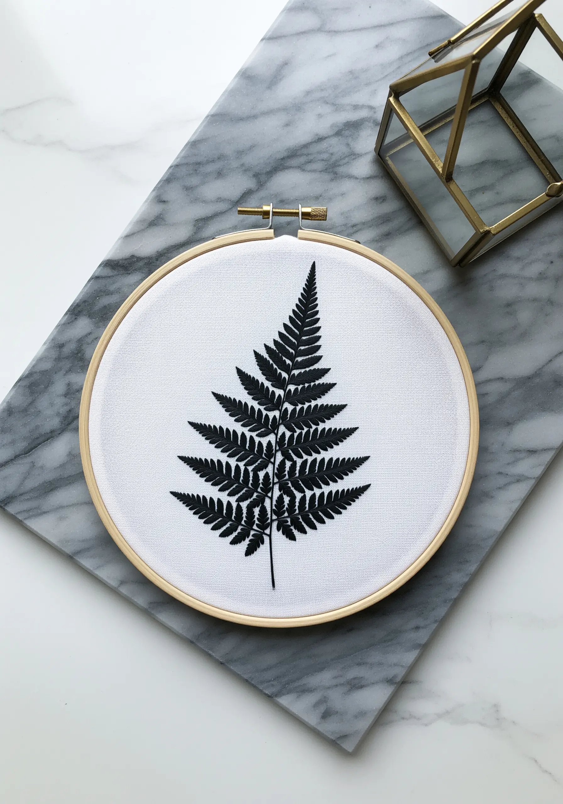 A detailed black fern silhouette embroidered on white fabric in a wooden hoop.