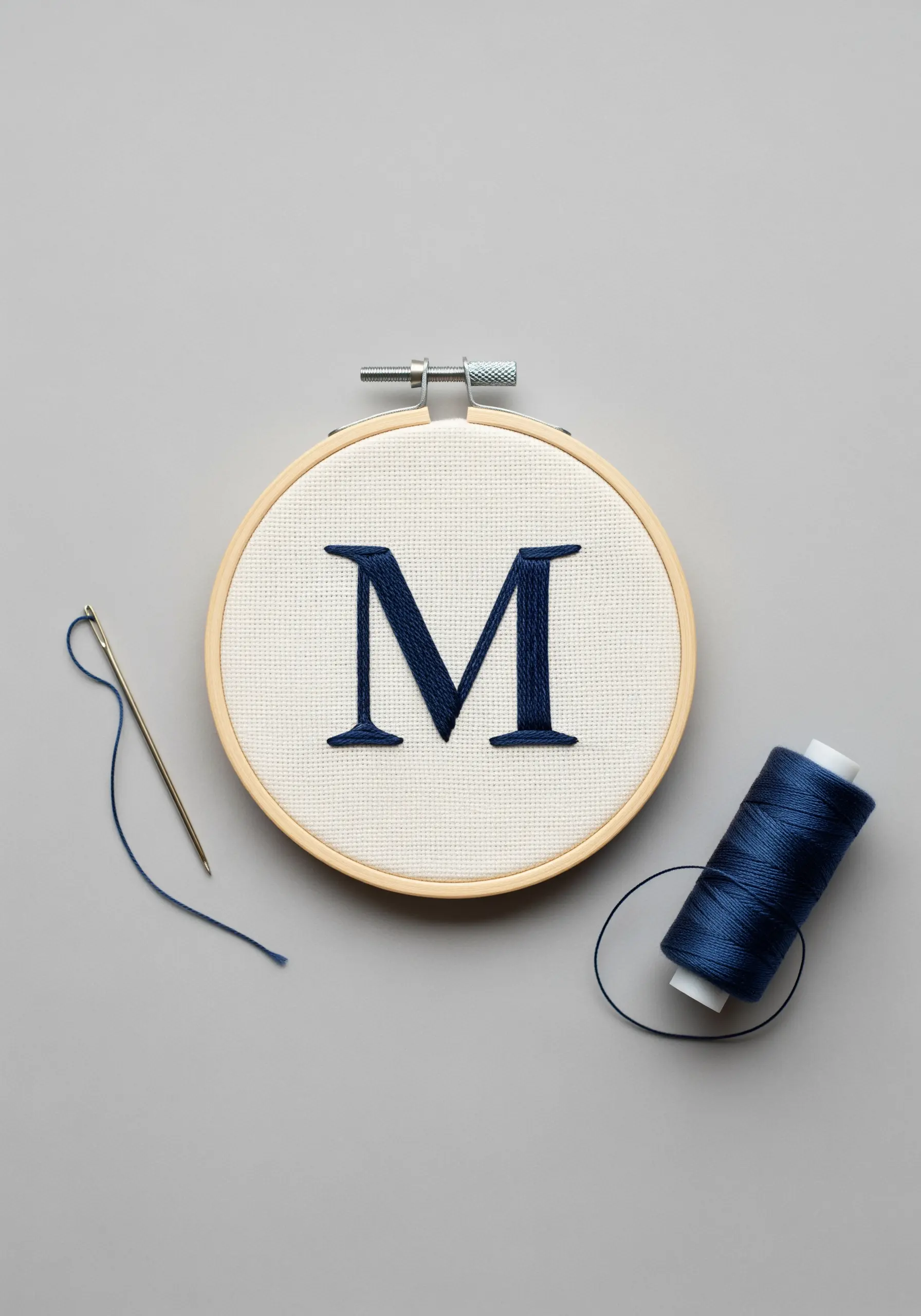 A bold, navy blue satin-stitched letter 'M' in a small wooden embroidery hoop.