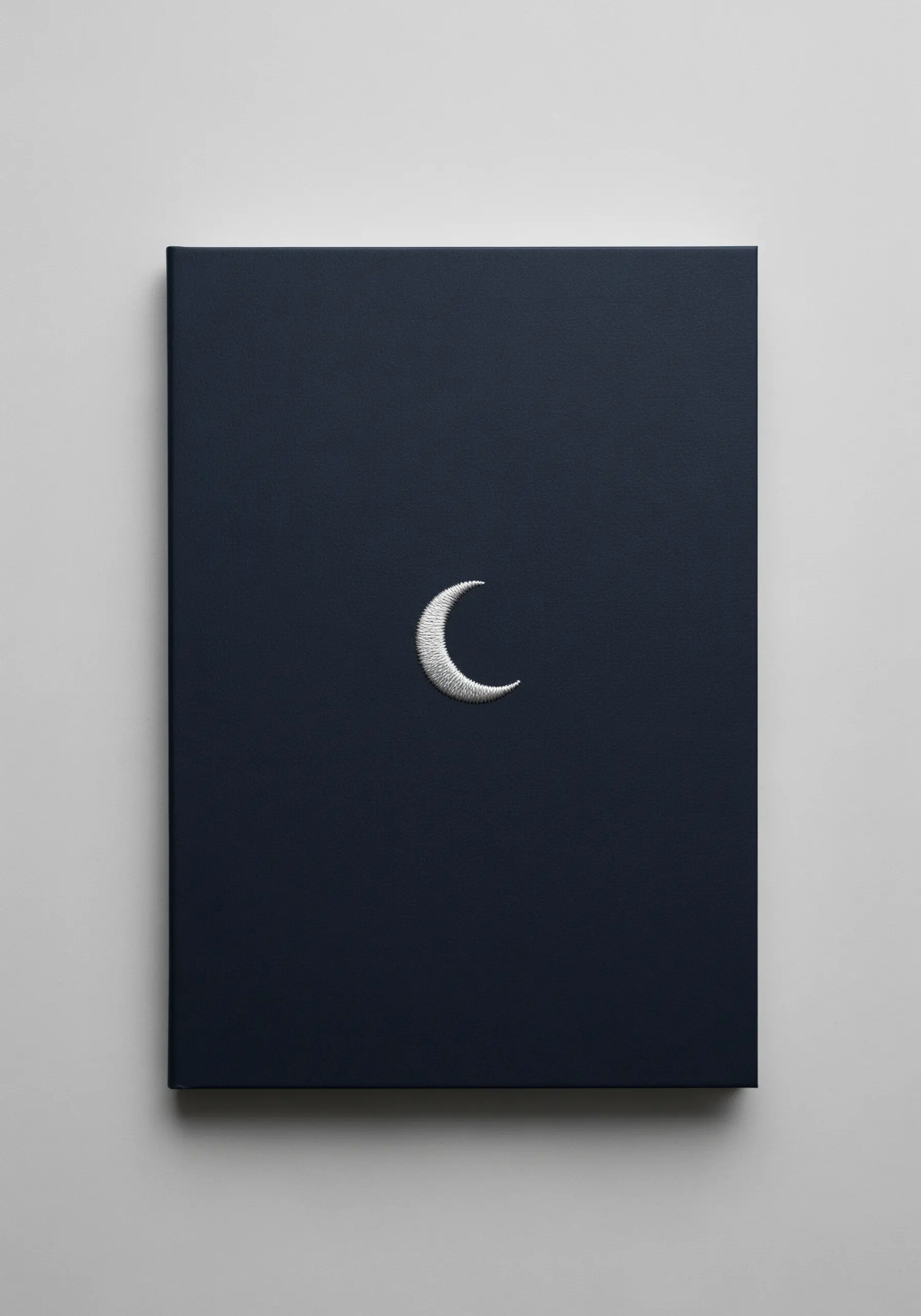 A tiny, sharp crescent moon embroidered in white thread on a dark blue notebook.