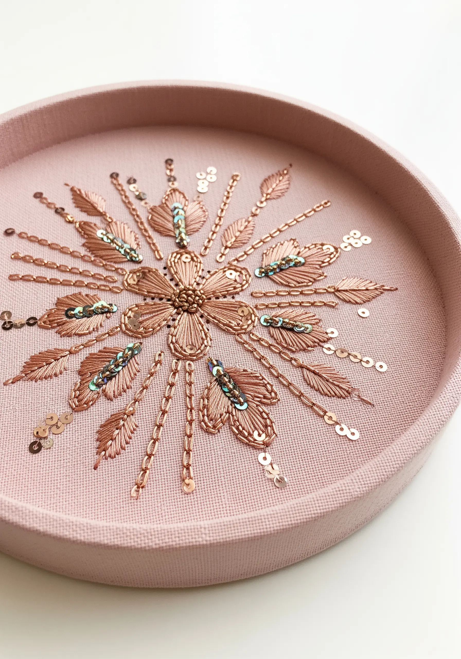 A floral mandala in copper metallic thread with sequin details in a pink frame.