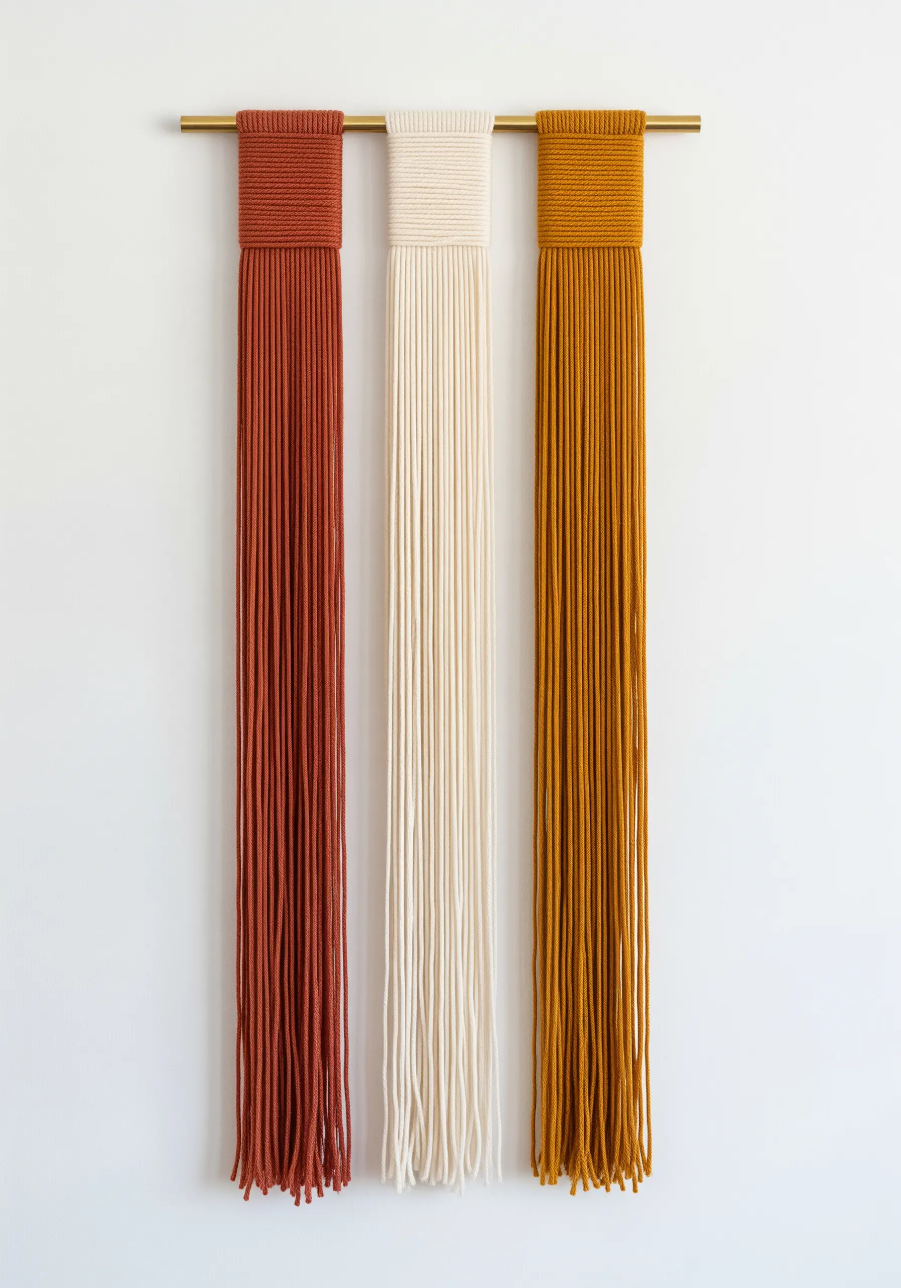 Three minimalist yarn panels in rust, cream, and mustard yellow hanging from a gold rod.