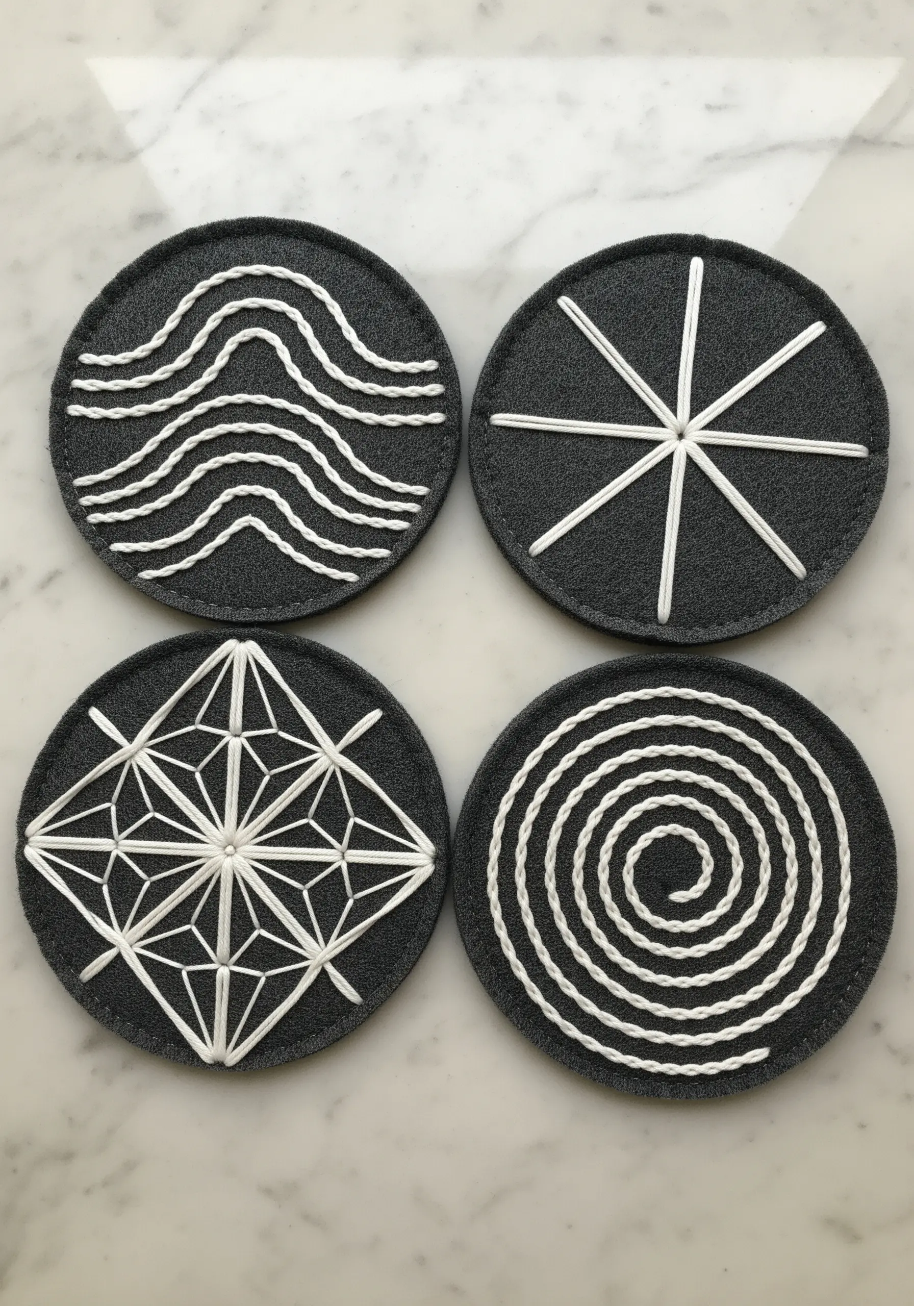 Four dark gray felt coasters, each embroidered with a different white geometric design.