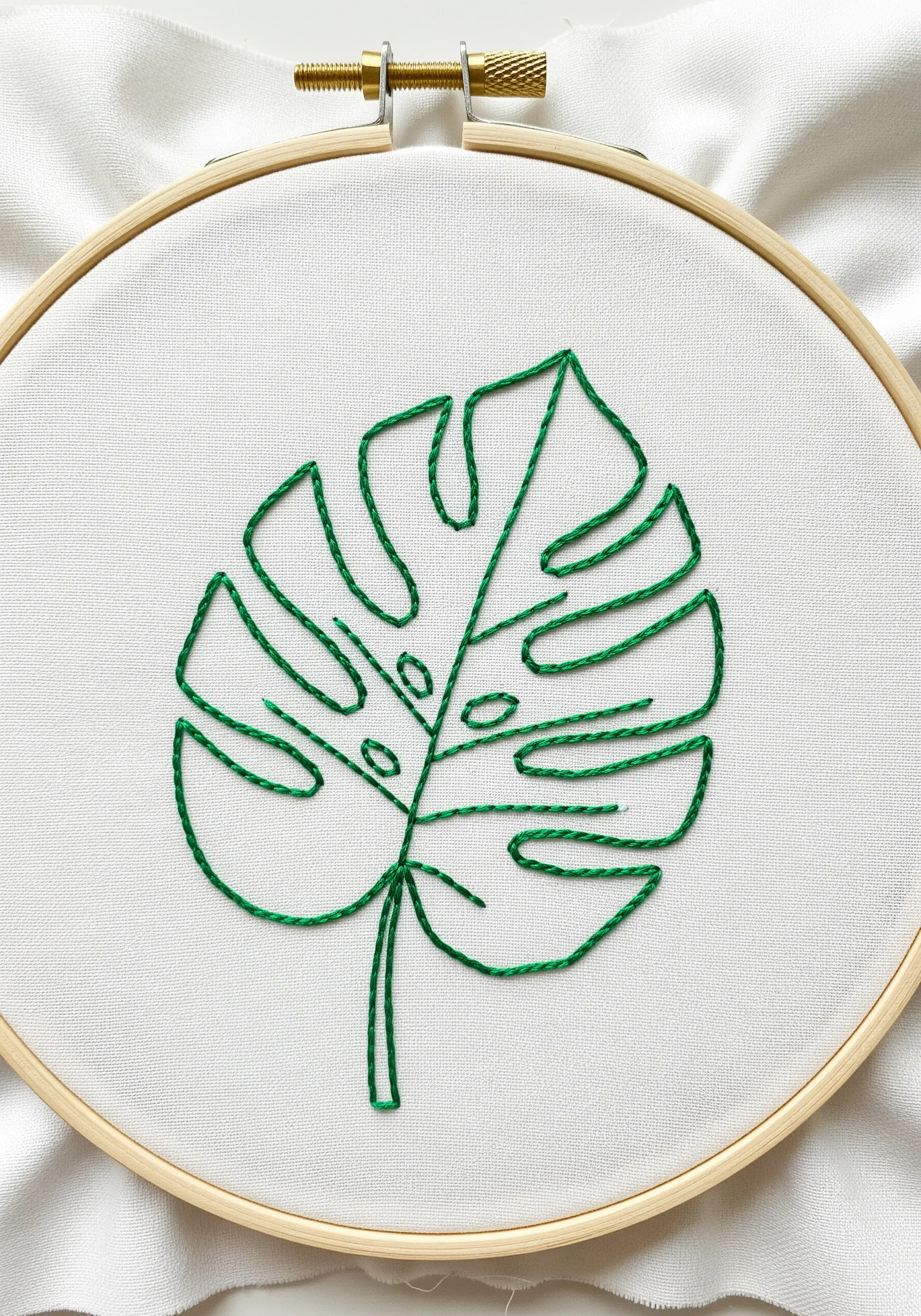 Minimalist line art embroidery of a green Monstera leaf on white fabric.