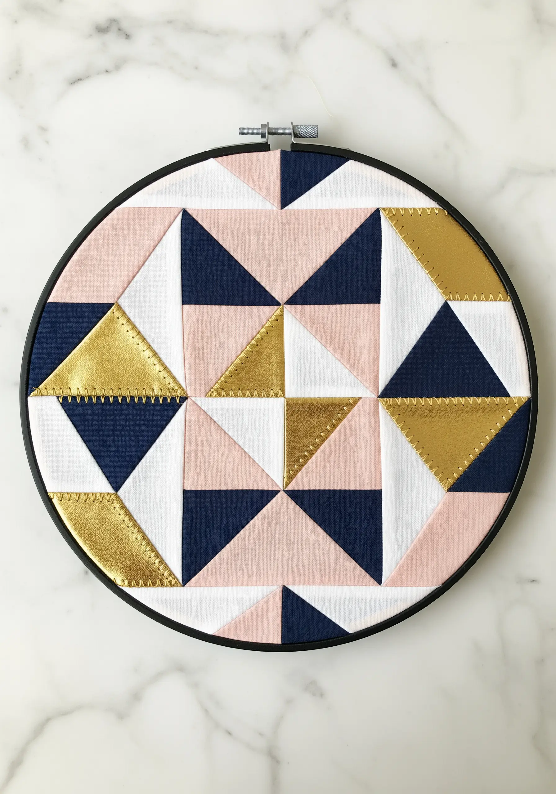 Geometric patchwork of pink, navy, white, and gold triangles in a hoop.