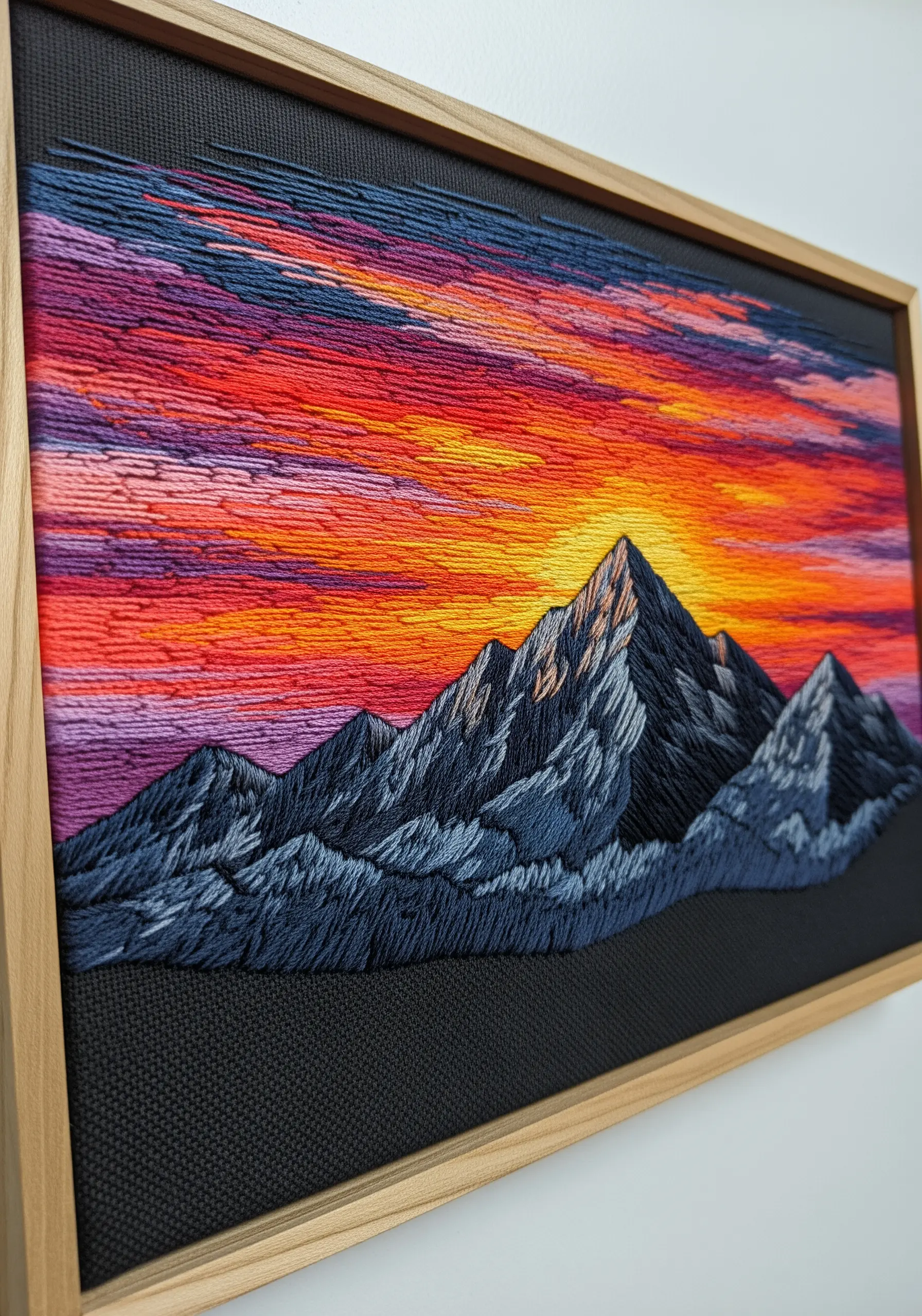 Framed thread painting of a mountain range against a vibrant, stitched sunset