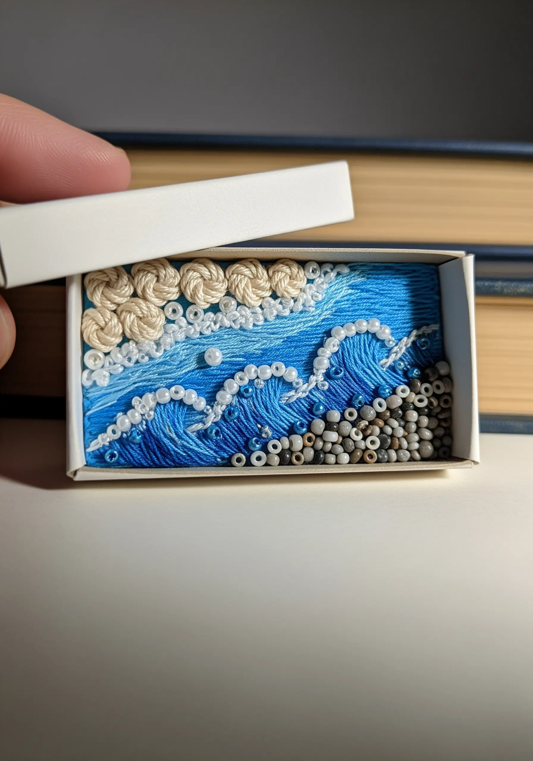 A miniature embroidered ocean scene inside a matchbox, with beaded waves and a pebbled shore.