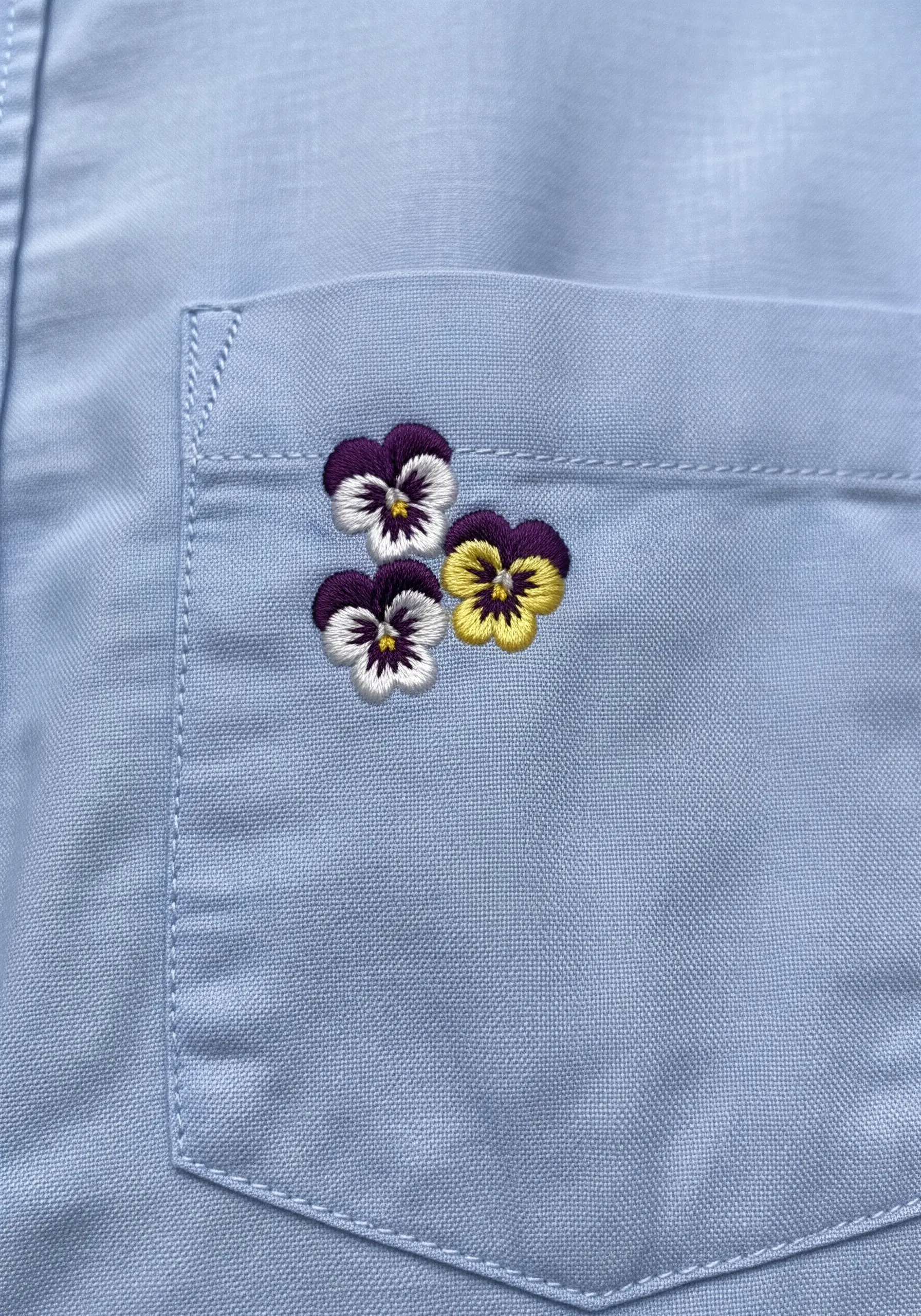 Three small, detailed pansies embroidered on the corner of a light blue shirt pocket.