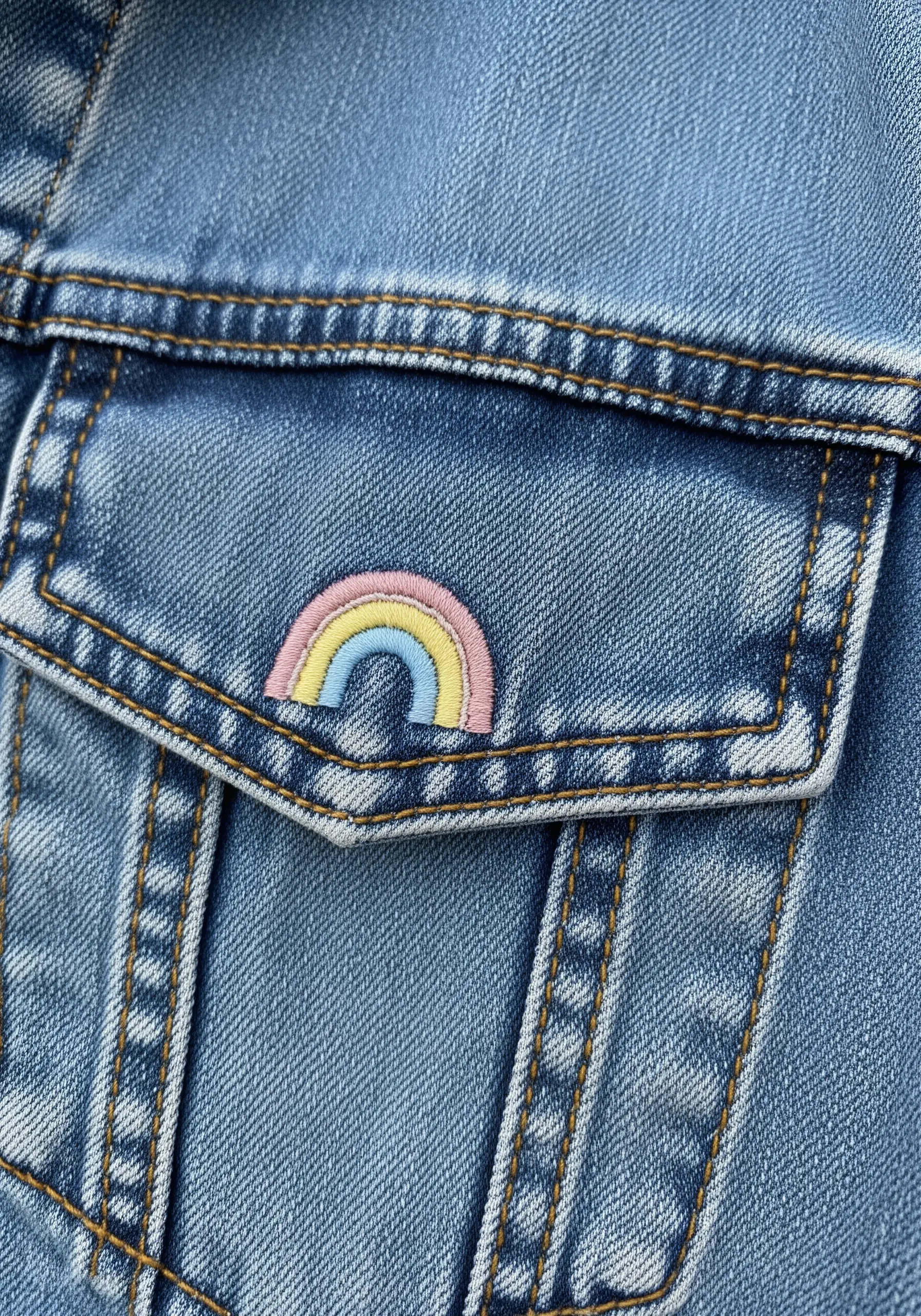 Small pastel rainbow embroidered on the pocket of a light blue denim jacket.