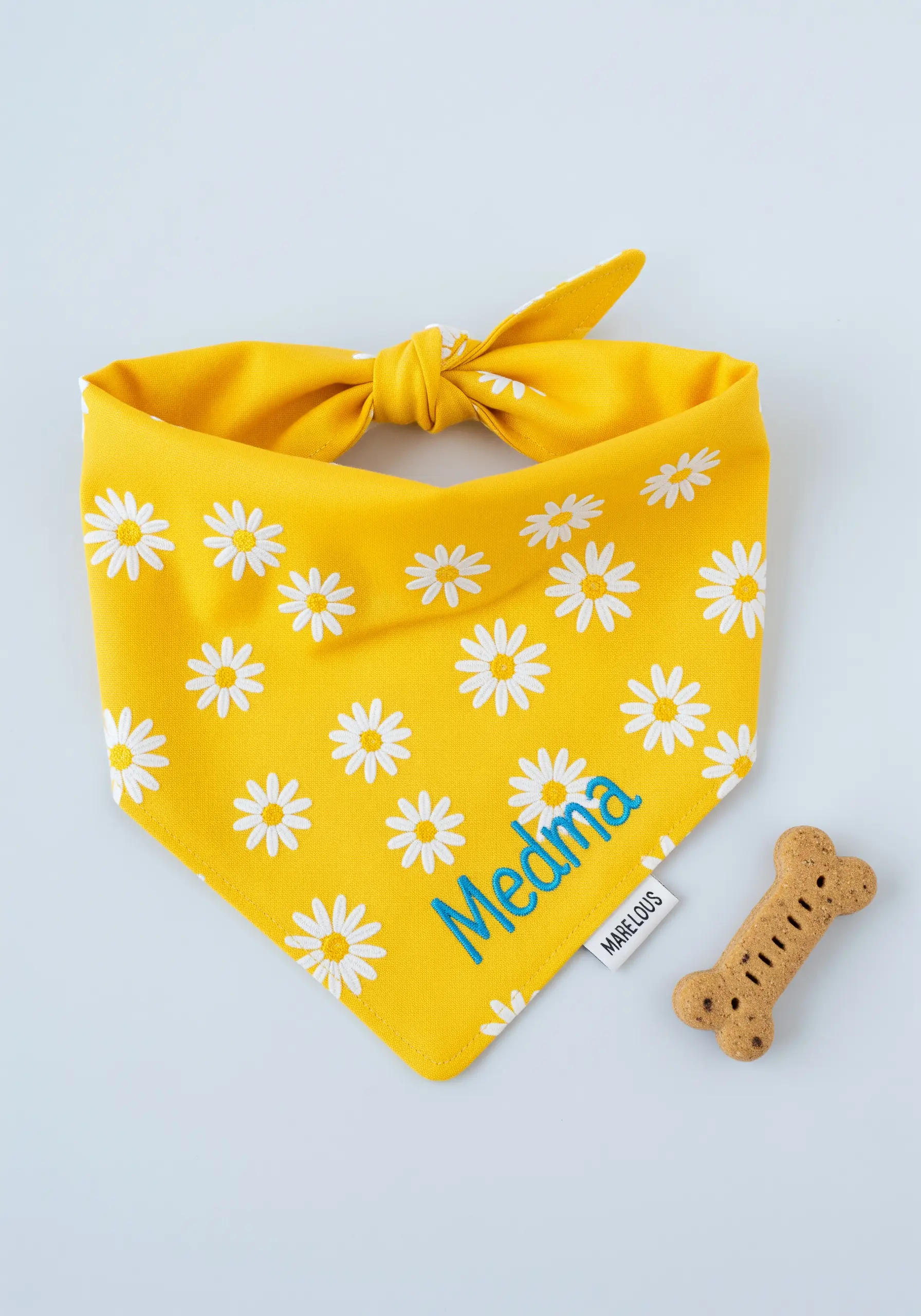 A bright yellow dog bandana with a daisy pattern, personalized with the name 'Medma' in blue thread.
