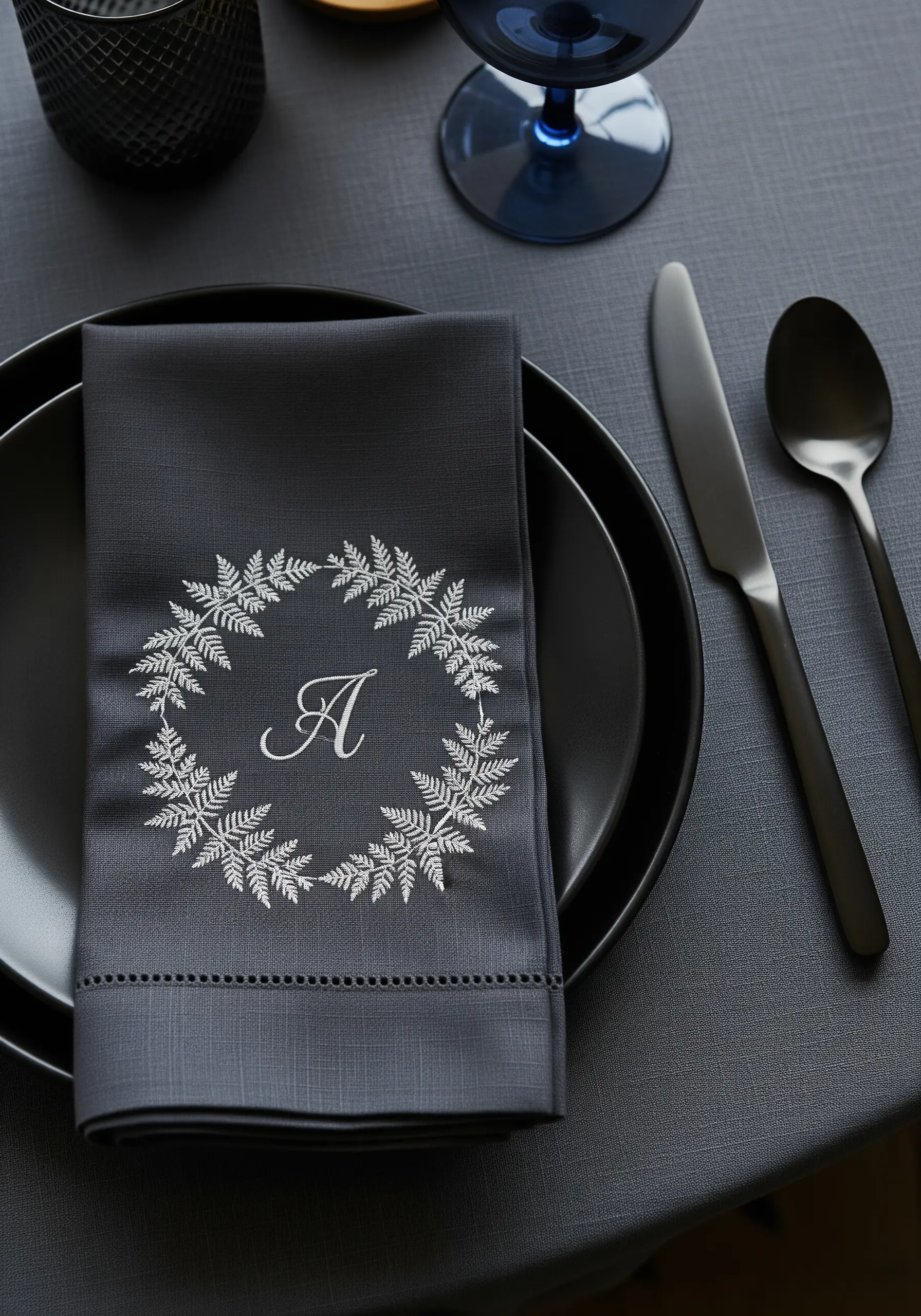 An elegant silver monogram 'A' inside a fern wreath embroidered on a dark grey linen napkin.
