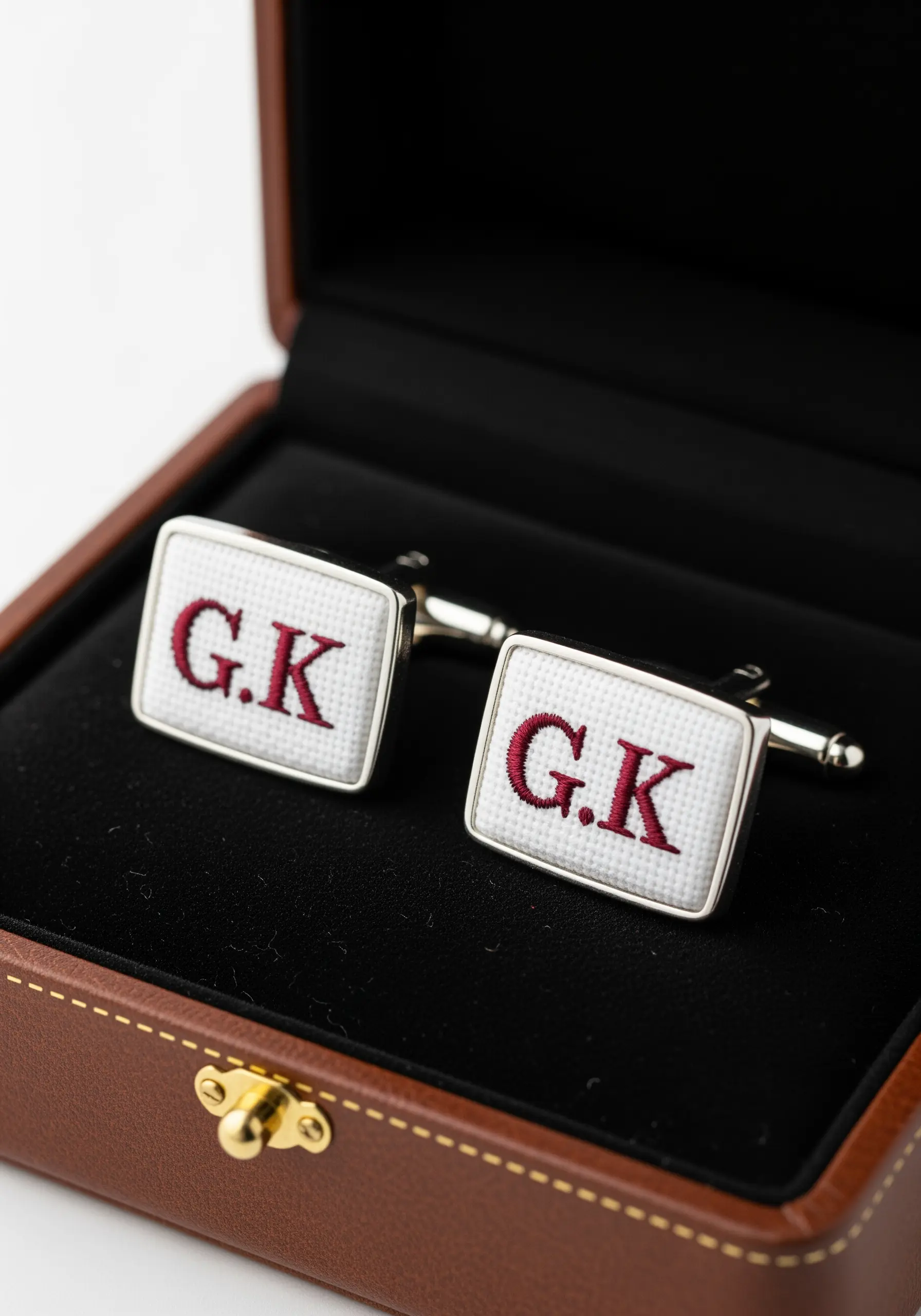 A pair of cufflinks with the initials 'G.K.' embroidered in red cross-stitch on white Aida cloth.