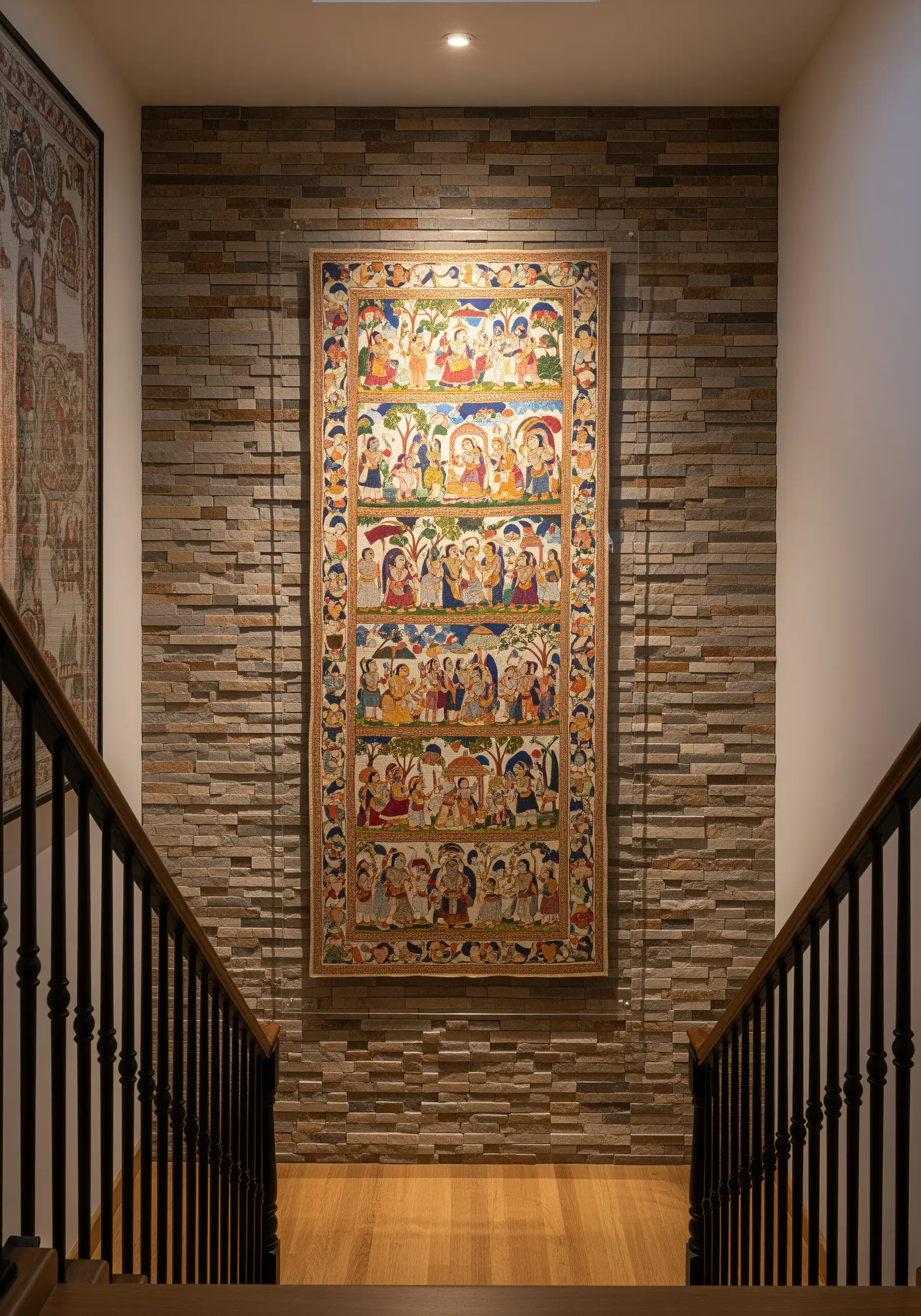 A tall, narrative Pichwai-style embroidered panel hanging on a stone wall in a stairwell.