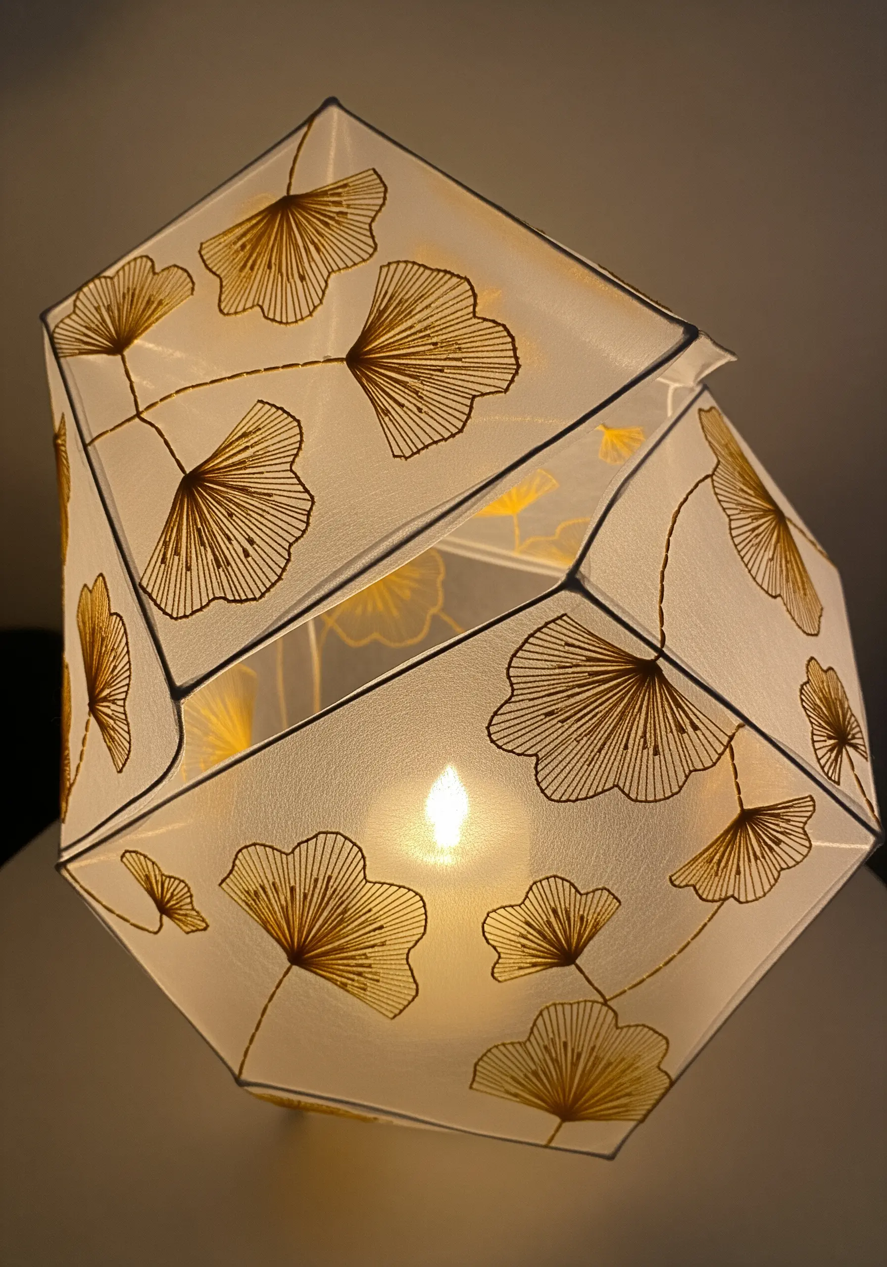 A geometric paper lantern illuminated from within, featuring embroidered ginkgo leaves.