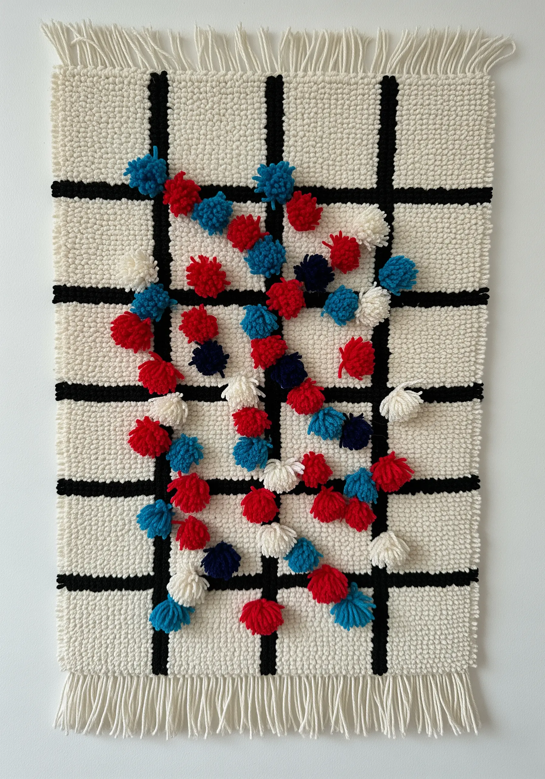 A woven wall hanging with a black and white grid, decorated with scattered red, white, and blue pom-poms
