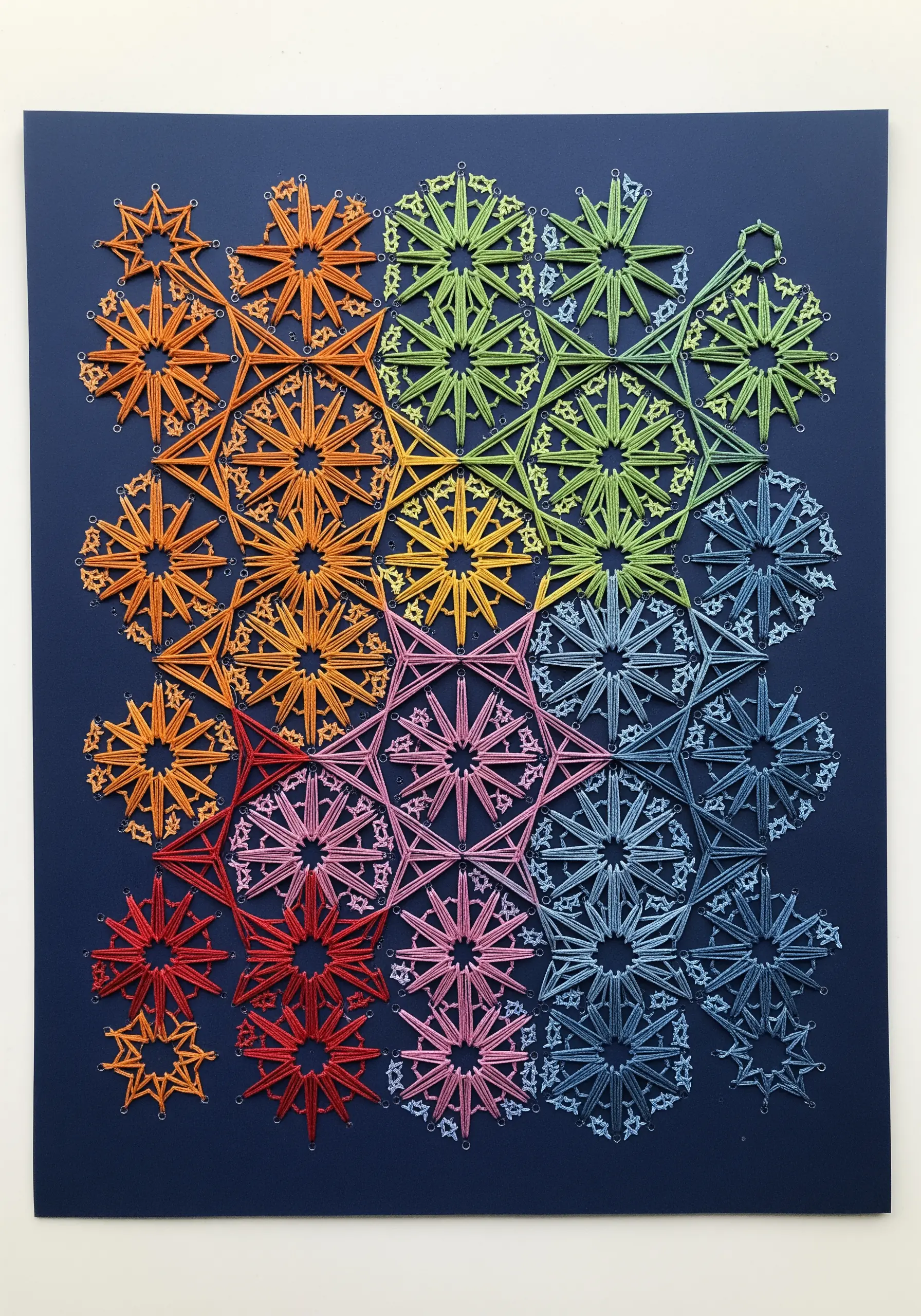 Intricate geometric starburst patterns stitched in a rainbow gradient on dark blue cardstock.