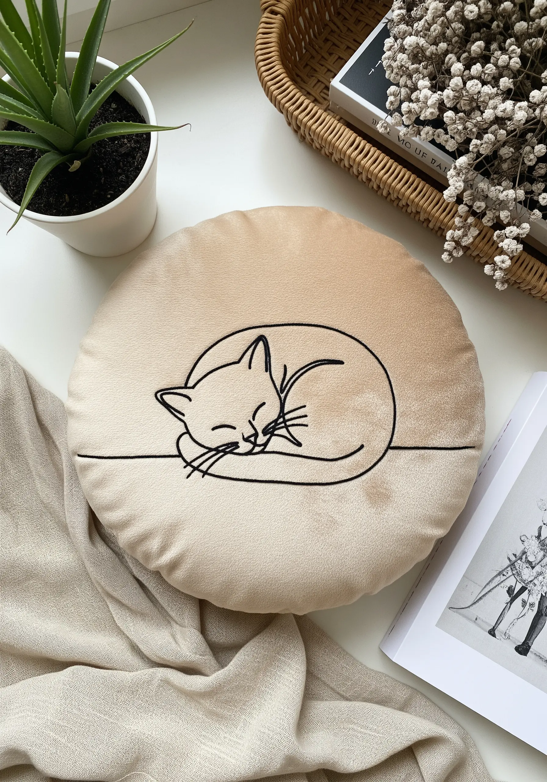 Line art of a sleeping cat embroidered in black on a round, plush beige cushion.