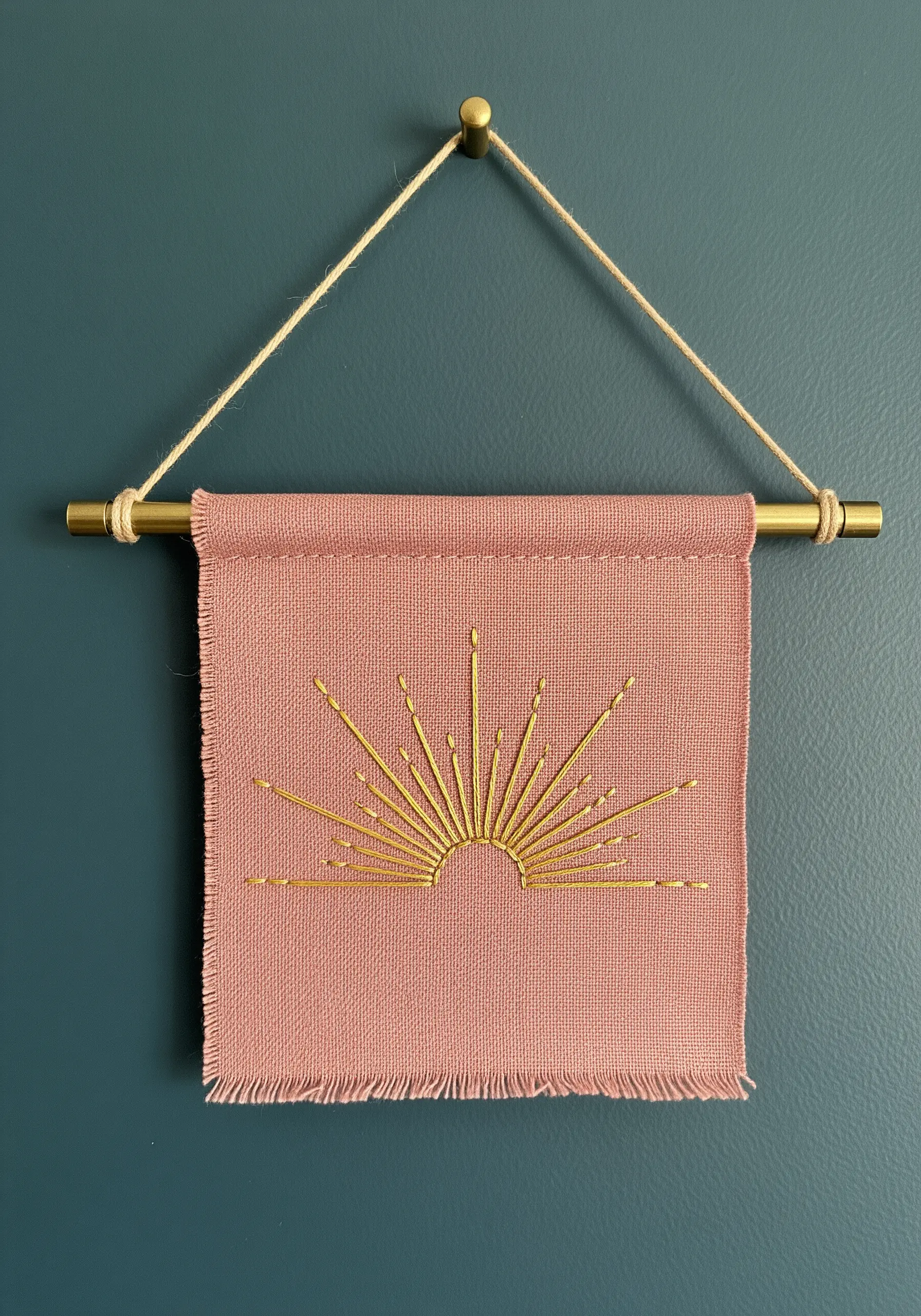 A metallic gold thread sunburst embroidered on a pink linen wall hanging with a frayed edge.