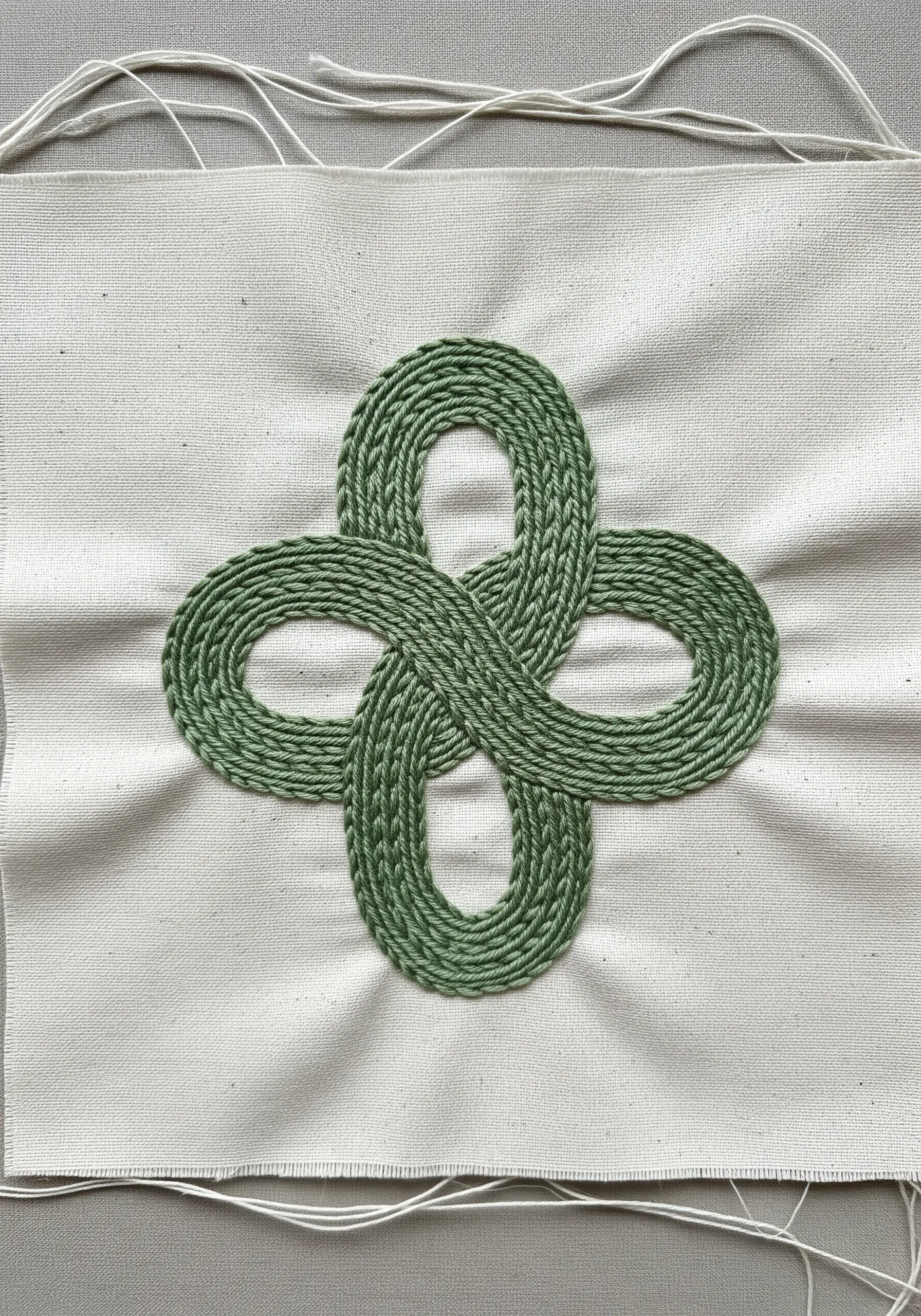 A green, interwoven Celtic knot with a raised, corded texture on white fabric.