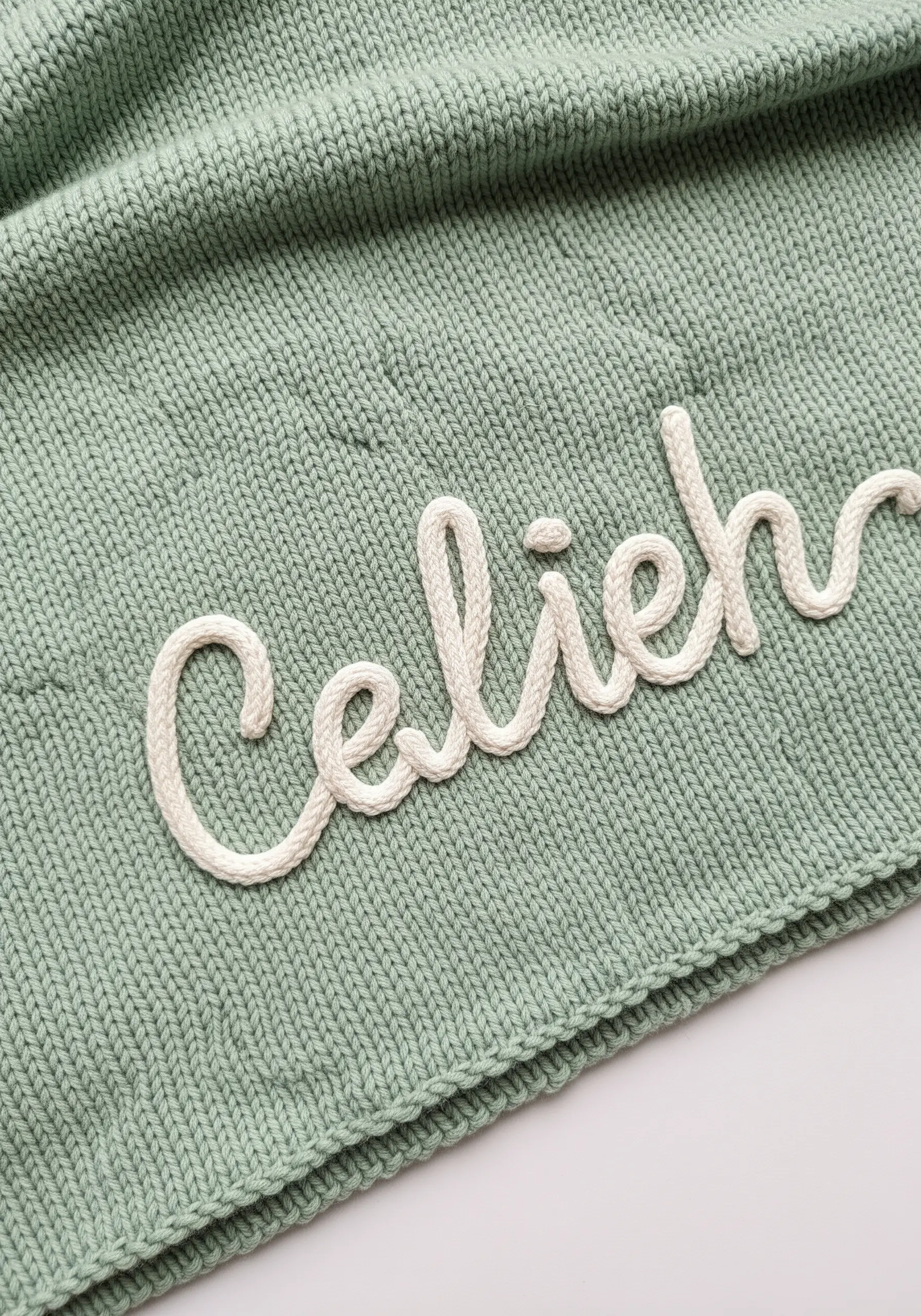 A baby's name embroidered in raised white chain stitch on a sage green knit blanket.