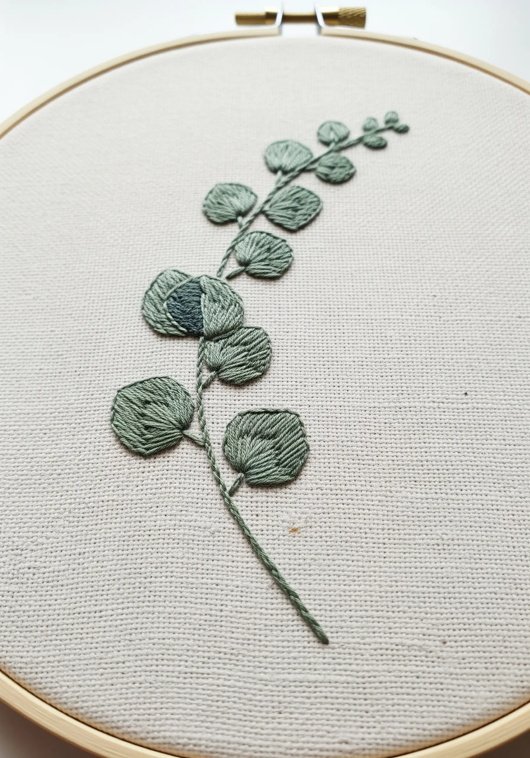 An embroidered eucalyptus branch with satin-stitched leaves on neutral fabric.