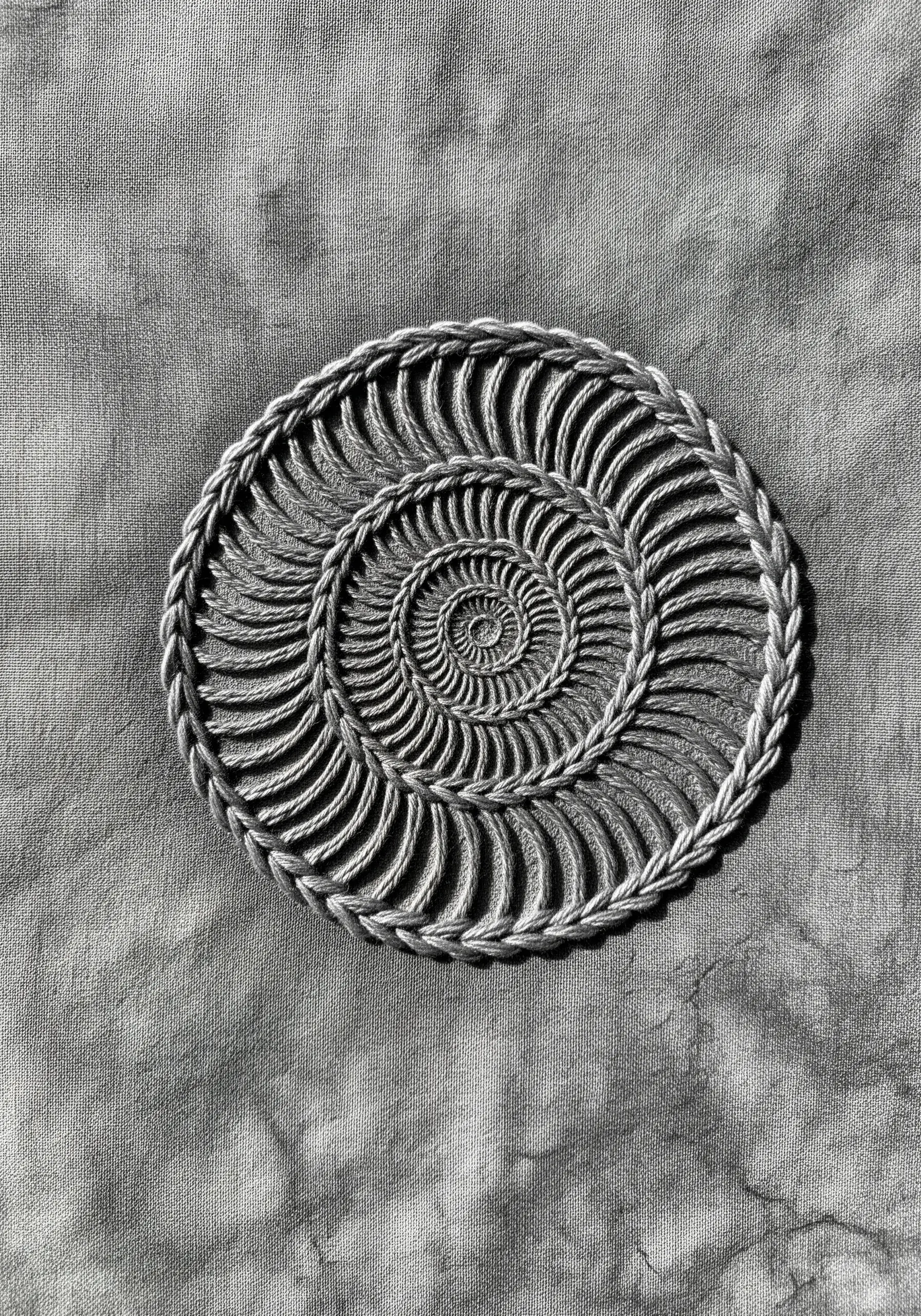 An embroidered ammonite fossil spiral on gray fabric using a raised rope-like stitch.