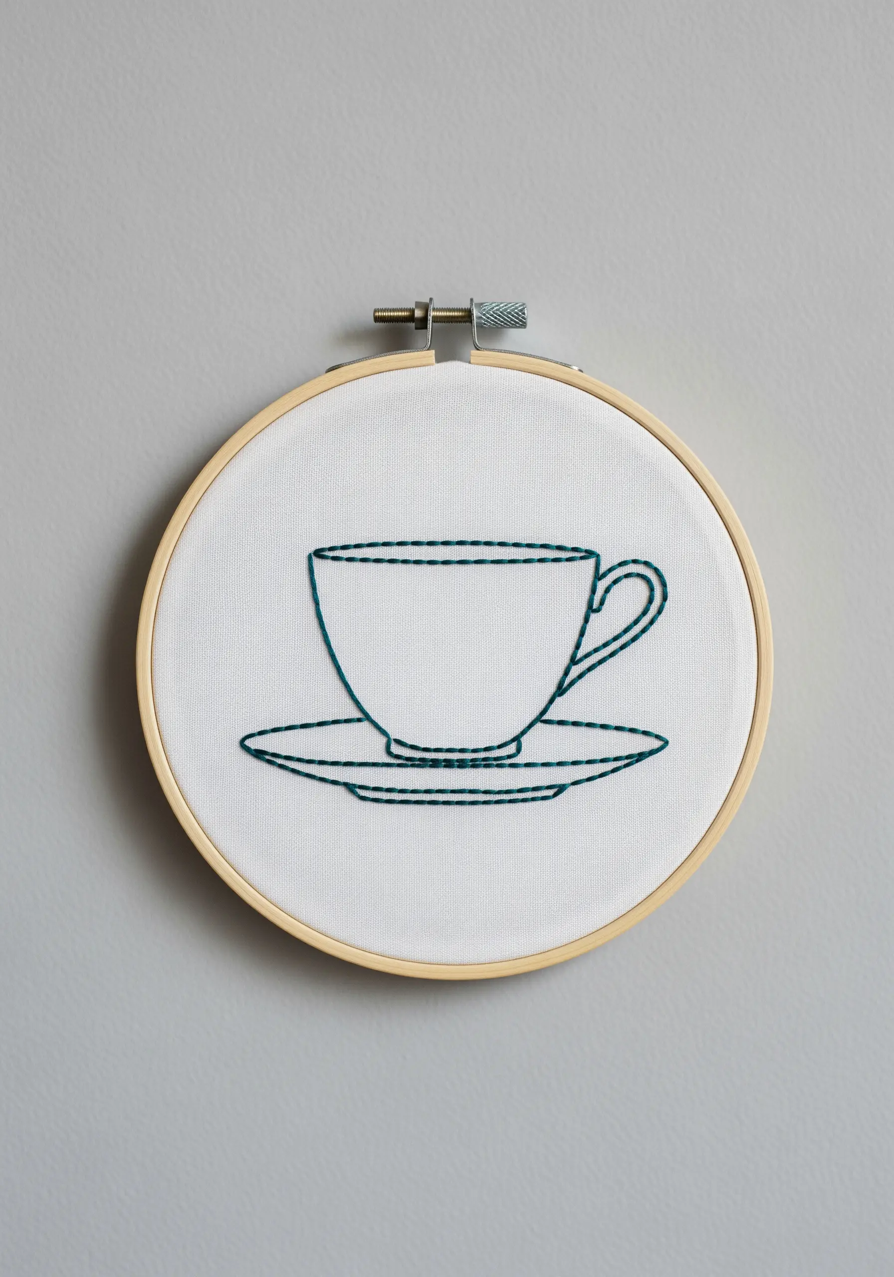 Minimalist outline of a teacup and saucer in dark teal thread on white fabric.